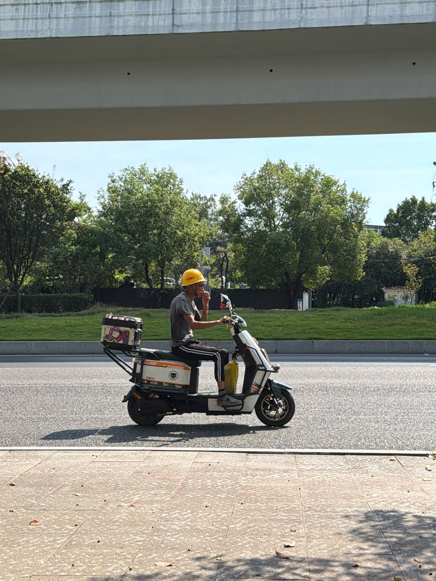 A person wearing a yellow hard hat rides a white and green electric scooter on a paved road in Jiangjin, China. The scooter has a black storage box on the back with colorful cartoon stickers. The rider is wearing a dark gray t-shirt and black pants with white stripes. The rider is holding something to their mouth. Behind the scooter, there is a grassy area with trees and a fence, and in the background, there is a building. Above the scene, a concrete overpass is visible. The ground in the foreground is paved with large stone tiles, and scattered leaves are visible.