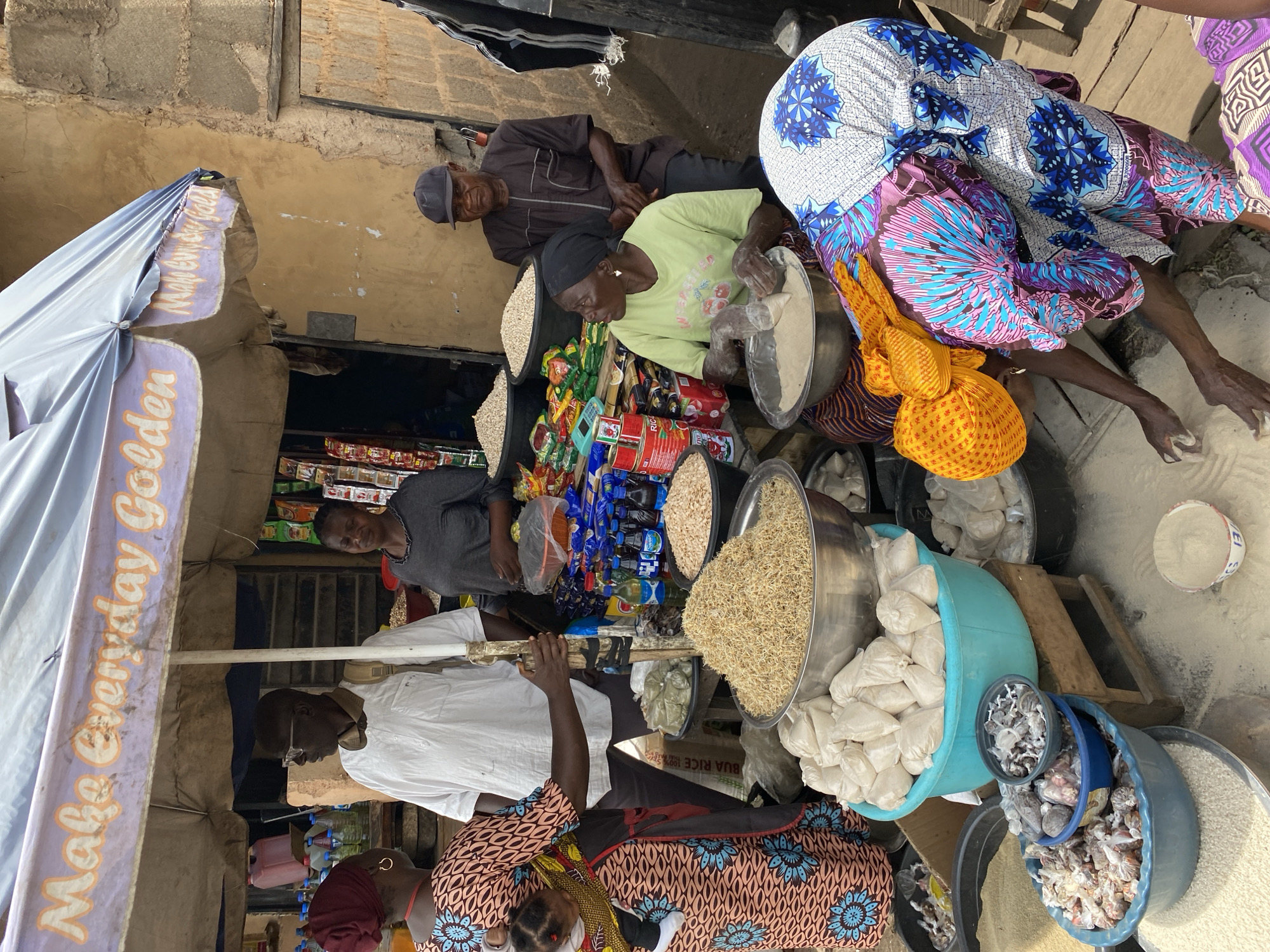 In Bukuru, Nigeria, a bustling market scene unfolds under a large, partially unfurled umbrella advertising "Make Everyday Golden." Several individuals are engaged in commerce and daily activities. In the foreground, a woman wearing a vibrant, patterned dress with blue and pink floral motifs is bent over, her hands sifting powder on a wooden surface. She is also wearing a bright yellow headscarf. To her left, a turquoise basin overflows with small, white, cloth-wrapped packages, likely goods for sale. Another basin next to it holds a dark gray powder, with a smaller blue bowl on top containing what appears to be dried fish or small meats. A large silver bowl in the mid-ground is filled with a mound of dried, granular material, possibly grains or legumes. Behind this, another woman in a grey t-shirt and headscarf stands near a display of various packaged goods, including bottles of drink and canned items. To her left, a man in a white long-sleeved shirt and glasses, with a backpack slung over his shoulder, stands attentive. Further back, a man wearing a dark cap and a brown patterned shirt is seated, observing the surroundings. The environment suggests an outdoor market with stalls constructed from wood and concrete. The ground is covered in a fine, light-colored powder, possibly related to the goods being sold.