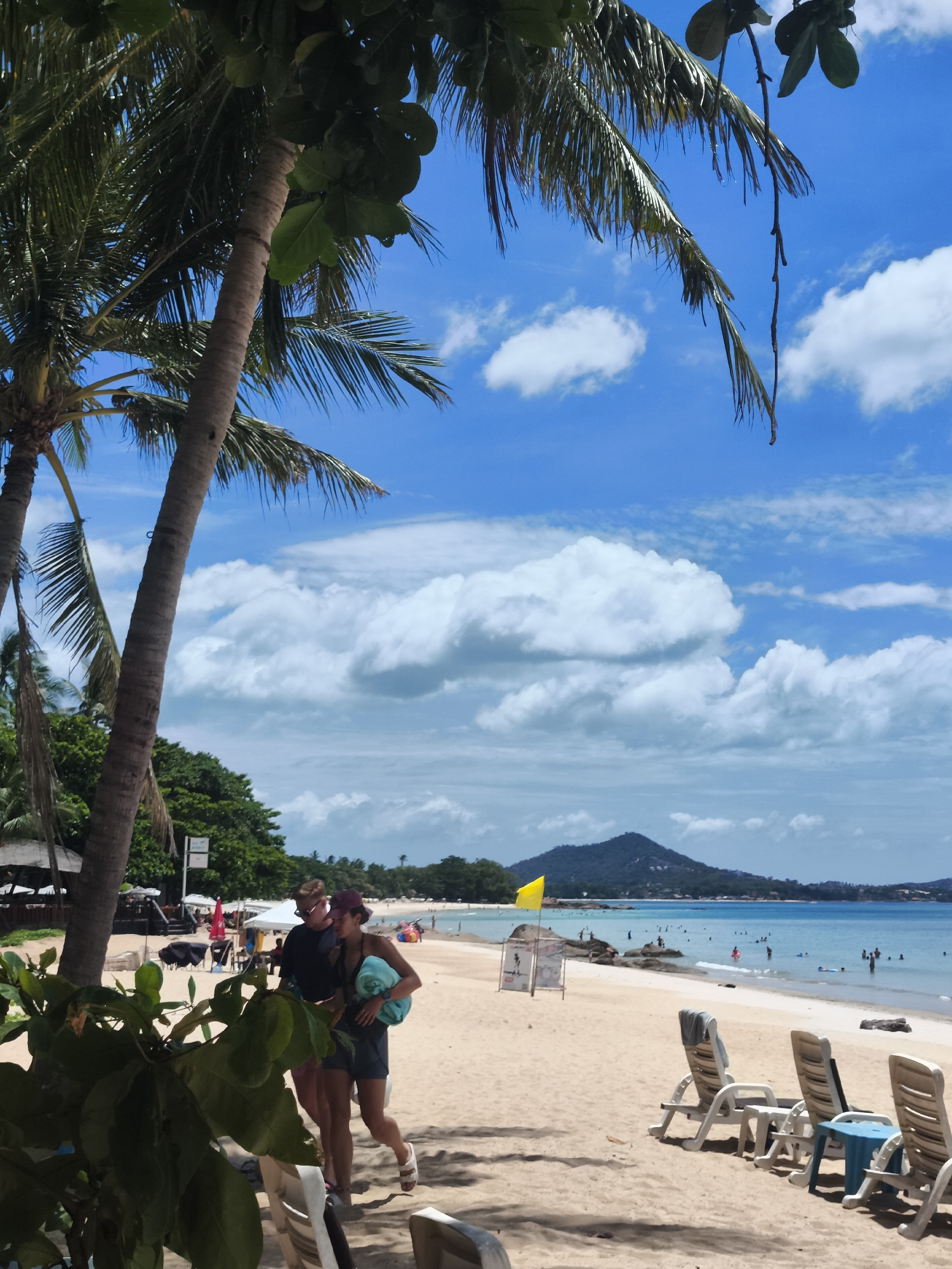 A daytime, eye-level, medium shot captures a bustling beach scene in the Ko Samui District of Thailand. In the foreground, two individuals, a man and a woman, walk side-by-side on the sand. The man, wearing sunglasses and a dark t-shirt, is on the left. The woman, wearing a dark top and shorts and a burgundy cap, is on the right, carrying a light blue towel. They are partially obscured by lush green foliage in the lower left corner.
Further down the beach, numerous beach chairs are set up on the sand, some occupied by people. The shoreline is lined with palm trees, and lush green vegetation extends inland. In the distance, people are swimming and relaxing in the clear blue ocean. A prominent yellow flag stands on a pole near the water's edge, accompanied by a sign. The horizon features a mountainous landscape under a bright blue sky dotted with fluffy white clouds. The overall atmosphere is lively and tropical.