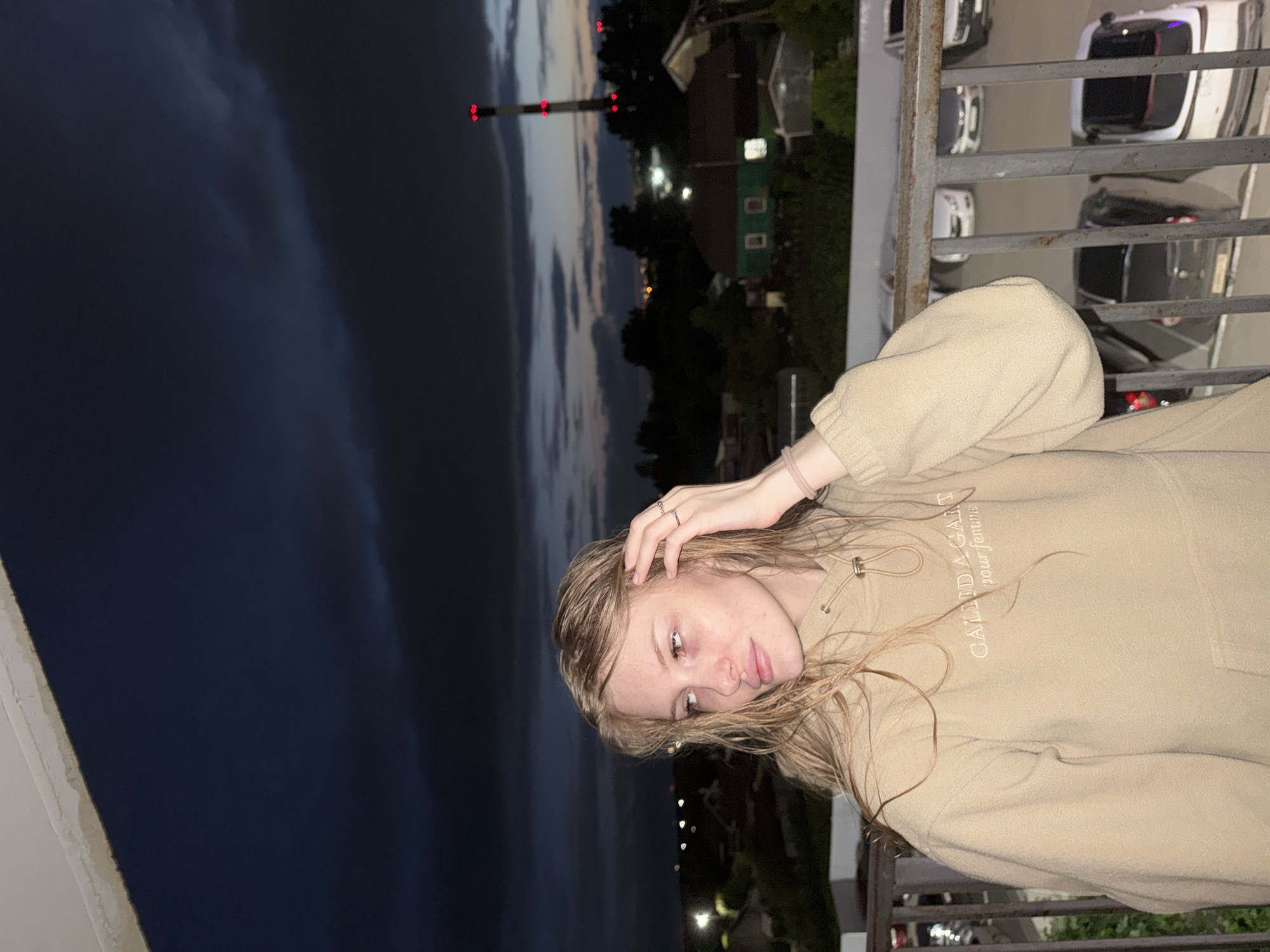 A young woman with blonde, wet hair stands on a balcony at dusk. She is wearing a tan hooded sweatshirt with text embroidered on the chest that reads "CALLIED A GARAGE pour femme." Her right hand is raised to her head, and she wears two rings on her finger. Behind her, a city skyline is visible, with houses, trees, and a tall smokestack with red lights. Cars are parked in a lot below the balcony. The sky is dark and cloudy.