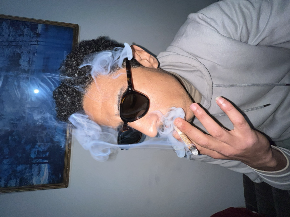The image depicts an indoor scene set in Tanger, Morocco. A male individual with dark, curly hair and brown skin, wearing dark tortoise-shell sunglasses and a light grey hooded sweatshirt over a beige turtleneck, is visible from the chest up, positioned towards the right side of the frame. He is holding a lit, hand-rolled cigarette (appearing to be a joint) between the thumb and forefinger of his right hand, positioned at his mouth. White smoke billows around his face, hair, and the cigarette. In the background, a light-colored wall is visible, with a framed painting hanging in the upper left portion. The painting features a dark blue, monochromatic landscape with trees and a body of water, illuminated by a bright, circular light source. A bright reflection point is present on the painting's surface. A dark, possibly upholstered object is partially visible in the lower left foreground.