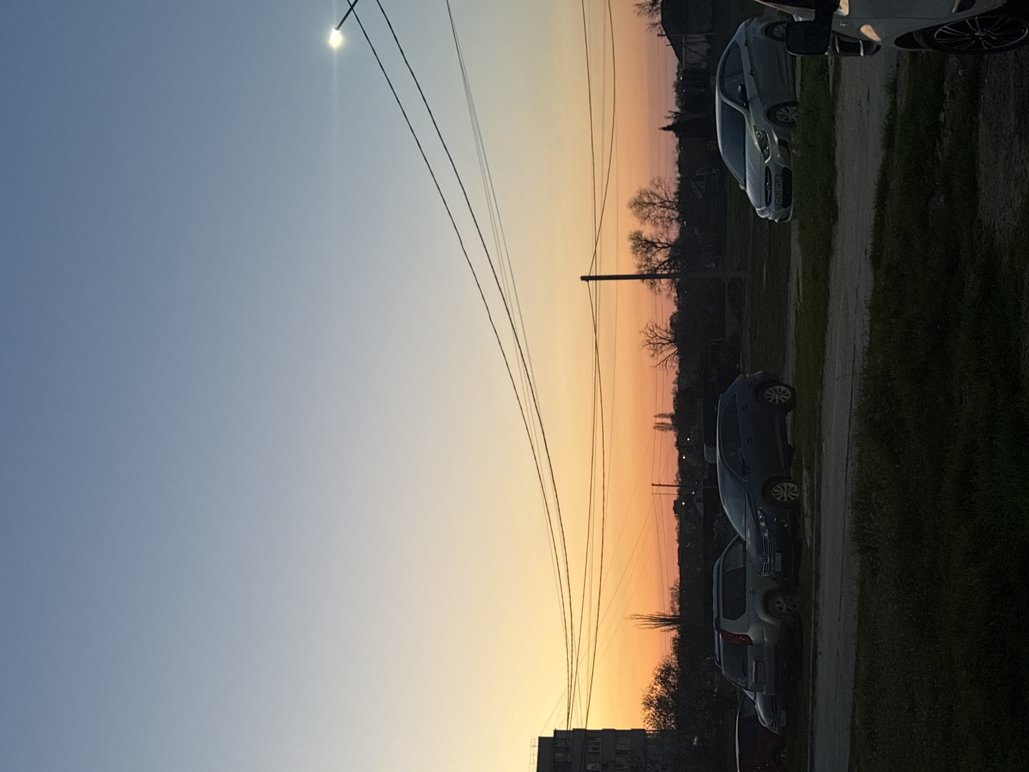 The image captures a street view in Chornomorskoe, Ukraine, at sunset. The sky transitions from a light blue to an orange-yellow hue near the horizon. Overhead power lines stretch across the sky. Parked cars line the right side of the image next to a strip of grassy area. A multi-story building is visible in the lower right corner, while silhouettes of trees and houses dot the horizon. The light suggests a low sun angle, casting long shadows.