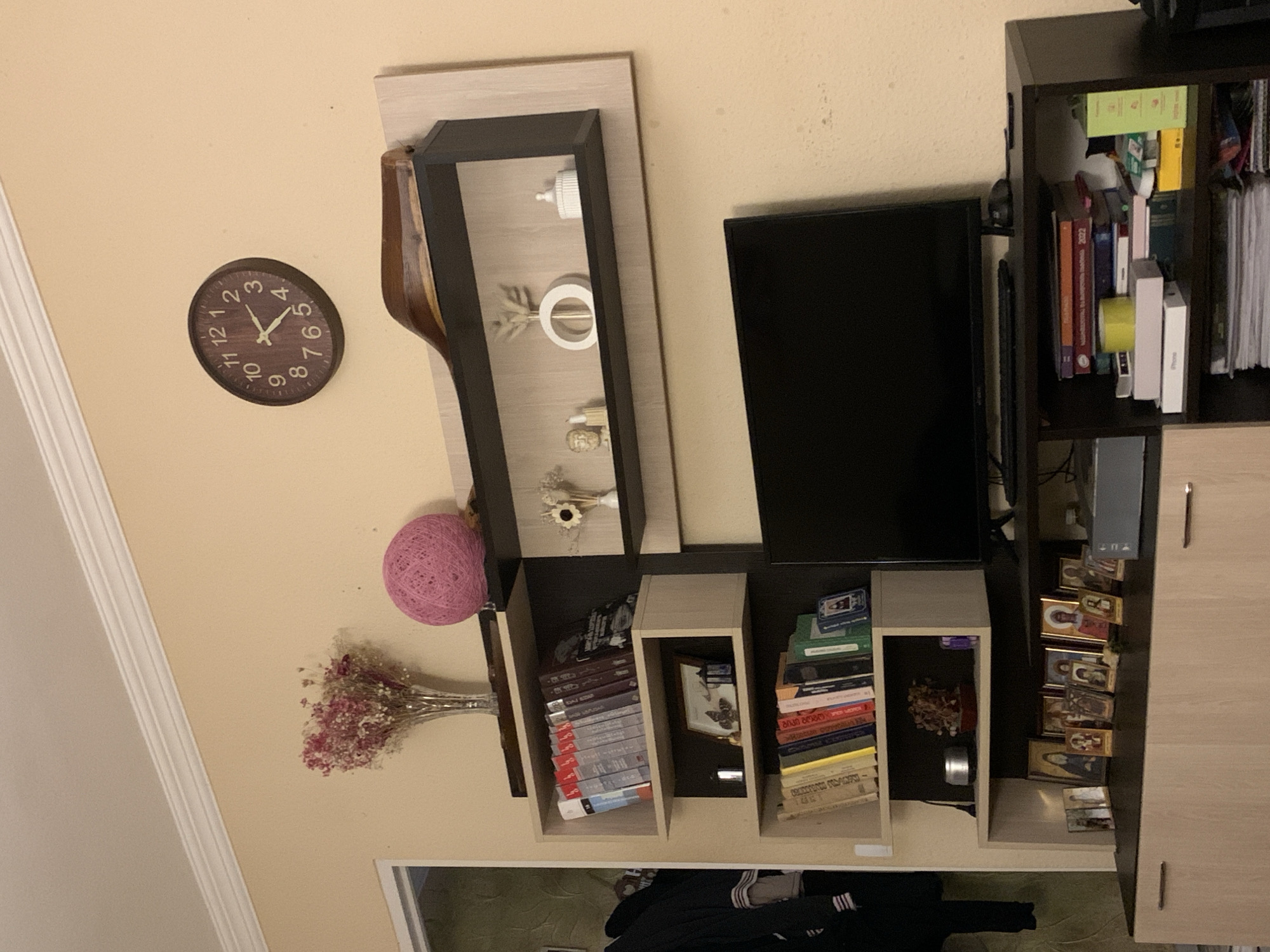 The image shows a living room interior with a wall-mounted entertainment unit. On the wall above the unit, a round, dark wood clock with white numbers and hands is visible. To the left of the clock, a vase of dried pink flowers sits on a shelf. A pink yarn ball is placed on a higher shelf.
The entertainment unit itself features several shelves. The top shelf holds a black-lined compartment with decorative items, including a small bust, a white vase with dried reeds, and a small white lidded container. Below this, a shelf is filled with a row of books. Another shelf below that has a framed picture with butterflies and a small blue lantern. The lowest shelf of this section is also filled with books.
To the right of the shelves, a black television is mounted. Beneath the television, a black entertainment stand holds more books and various other items on its shelves, including a yellow roll of tape and boxes. The bottom part of the image reveals a light-colored cabinet with drawers. On top of this cabinet, a collection of framed religious icons is displayed. The room has beige walls and white trim along the ceiling. The scene appears to be a home environment, possibly in an apartment.