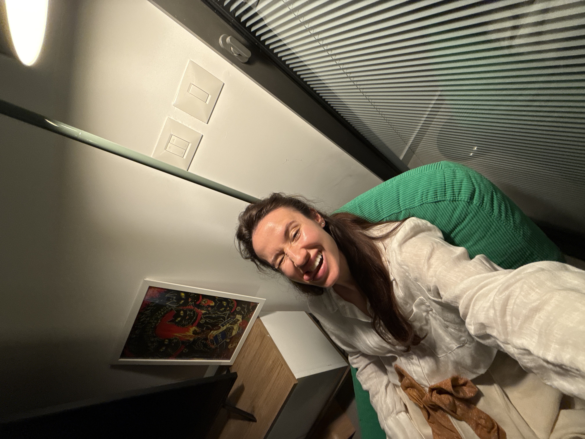 A low-angle indoor shot depicts a woman smiling and sticking out her tongue while sitting on a green corduroy-covered surface. She wears a light-colored linen shirt and pants. Behind her is a wall with light switches and a framed artwork. A light source illuminates the scene from the upper left. The background includes window blinds. There is no evidence to indicate the image was taken in Tel-Aviv, Israel.