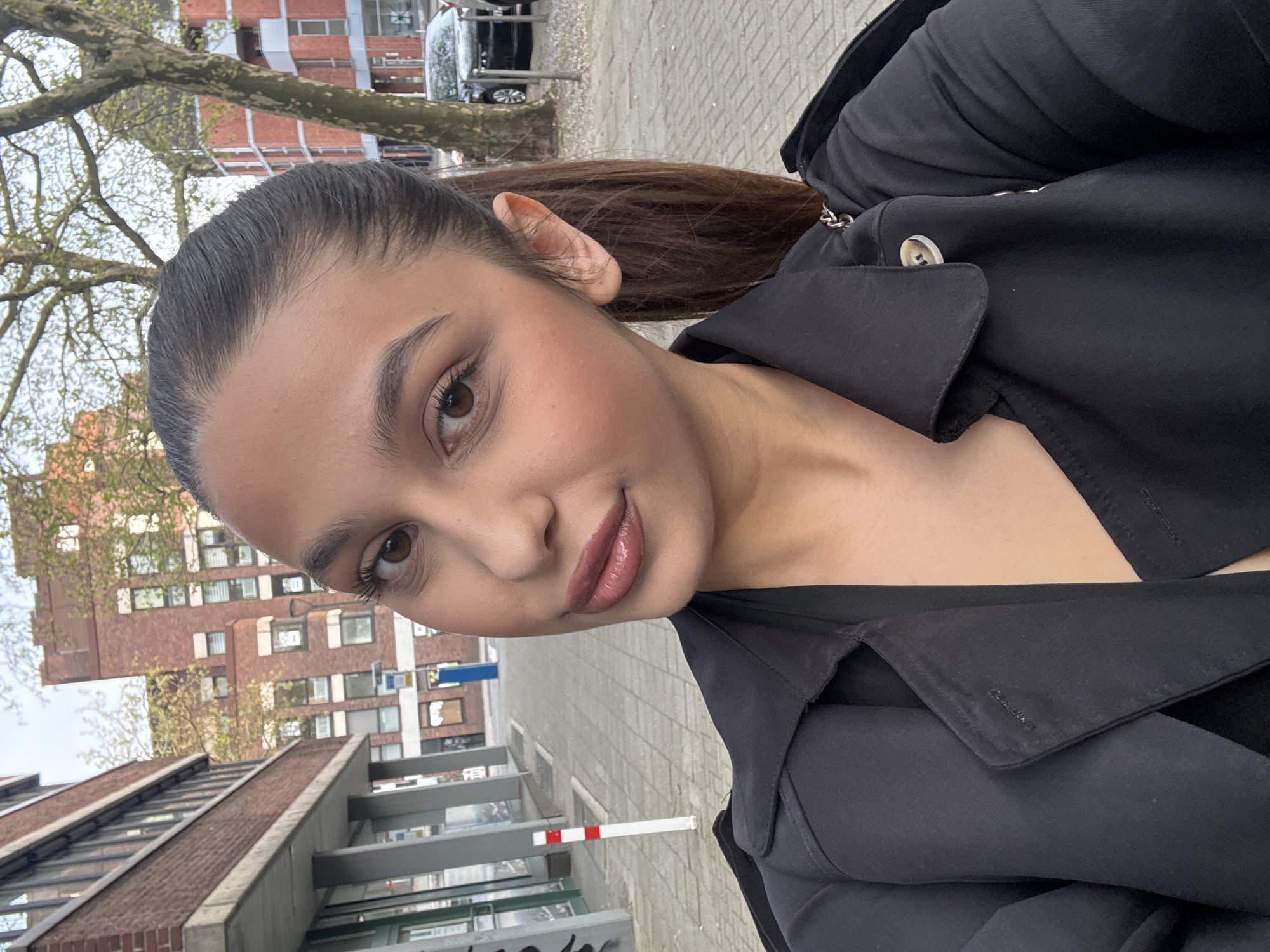A selfie of a woman with brown hair pulled back, wearing a black trench coat. She is standing on a tiled sidewalk in Mönchengladbach, Germany. In the background is a building made of brown brick. A tree with budding leaves is visible on the left. A vehicle is parked in the distance.