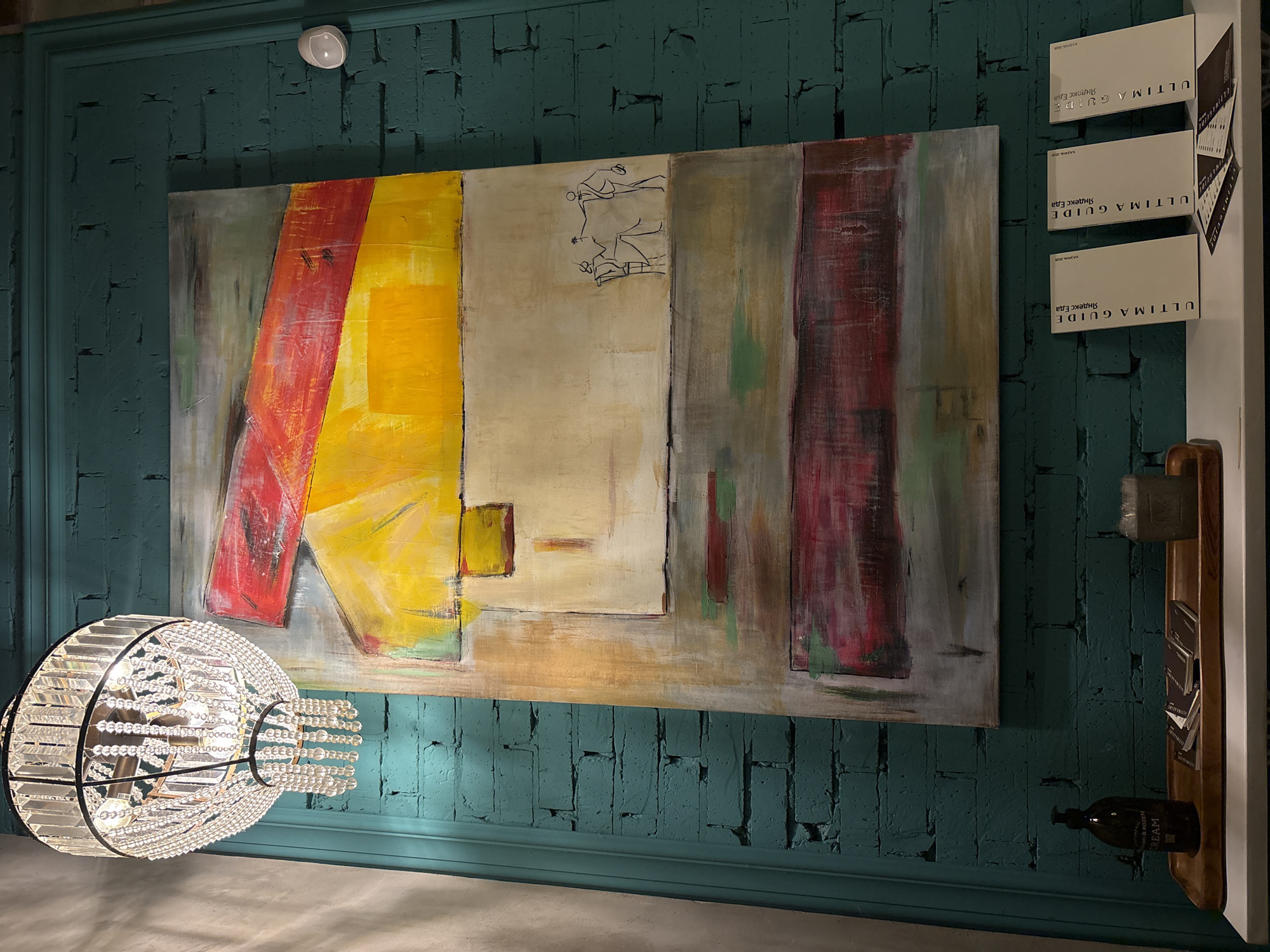 Here is a forensic description of the image:
**Scene:** Interior shot of a room with a textured teal wall, likely in Kazan, Russia, based on the text "КАЗАНЬ 2023" printed on the "Ultima Guide Яндекс Еда" brochures. The wall appears to be brick painted in teal, showing signs of wear and texture.
**Objects:**
* **Painting:** A large abstract painting hangs on the wall featuring bold blocks of red, yellow, and grey. The painting also includes a thin line drawing of two people.
* **Chandelier:** A decorative chandelier hangs in the lower-left corner of the frame. It is constructed of metal framing and adorned with crystal or glass beads.
* **Brochures:** Three rectangular cards stacked vertically on the right with the text "ULTIMA GUIDE Яндекс Еда" on them. They are likely restaurant guides.
* **Shelf:** A narrow wooden shelf is mounted on the wall to the right of the painting.
* **Objects on Shelf:** A small brush or duster rests on the top of the shelf. Below the brush, there appear to be more cards or brochures stacked vertically. A dark bottle, likely containing lotion, sits at the bottom of the shelf with text "CREAM" written on it and "K&ROZEN" printed above.
* **Light Fixture:** A small, white circular light fixture is mounted on the wall above the painting.
**Environment:** The room's ambiance appears dimly lit, casting shadows and emphasizing the texture of the wall. The color palette is dominated by teal, red, yellow, grey and tones of cream and brown.