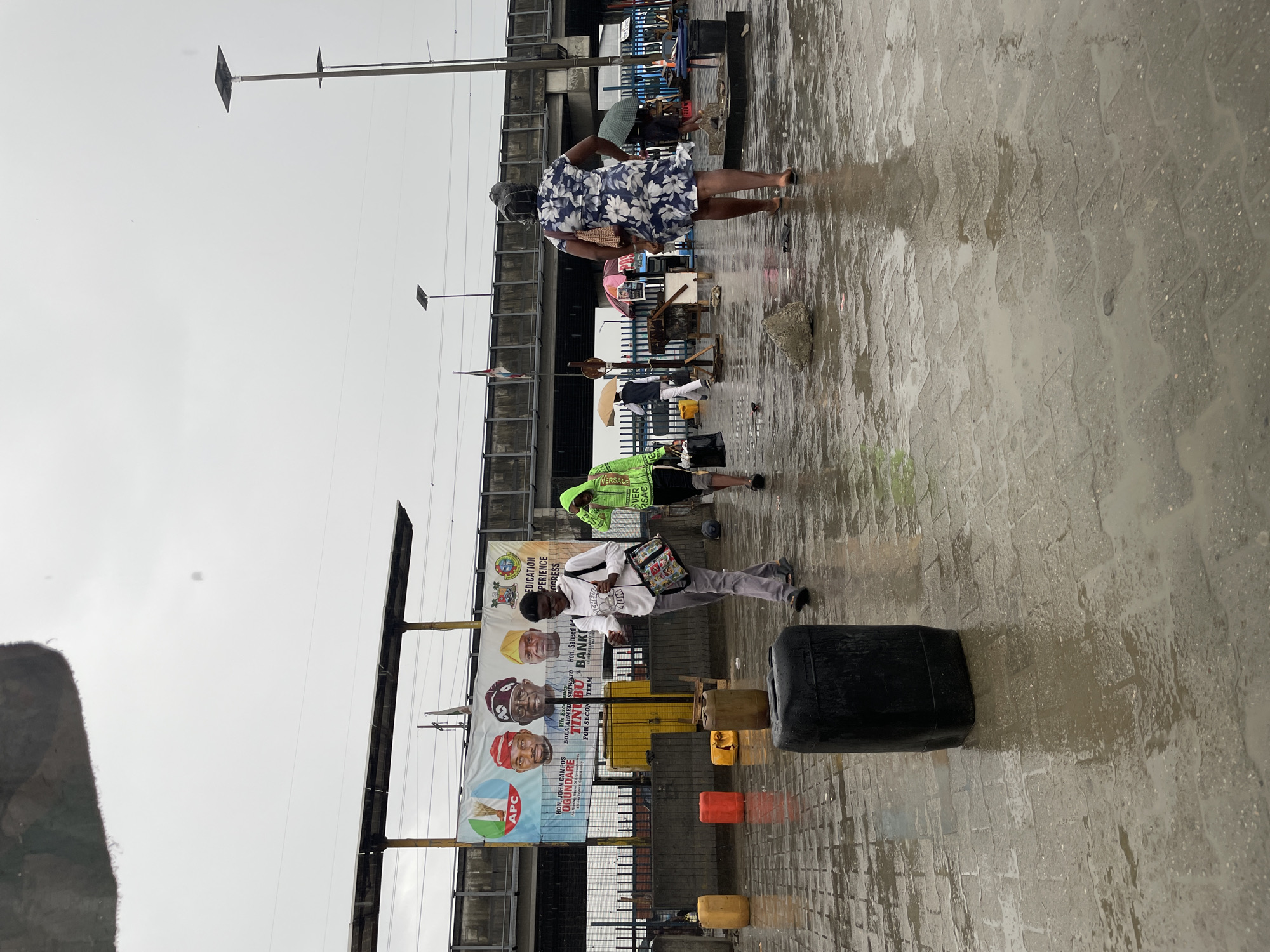 The image depicts a street scene in Lekki, Nigeria, on what appears to be a wet day. The ground is paved with cobblestones and covered in puddles. An elevated structure with electrical poles and wires runs parallel to the street. A large banner with political figures and the abbreviation "APC" (likely a political party) is visible. Several people are present: a woman in a floral dress, a man carrying a container, and another man wearing a bright green top. Objects include plastic containers, a black rectangular container, and street vendor setups. The people appear to be moving along the street.