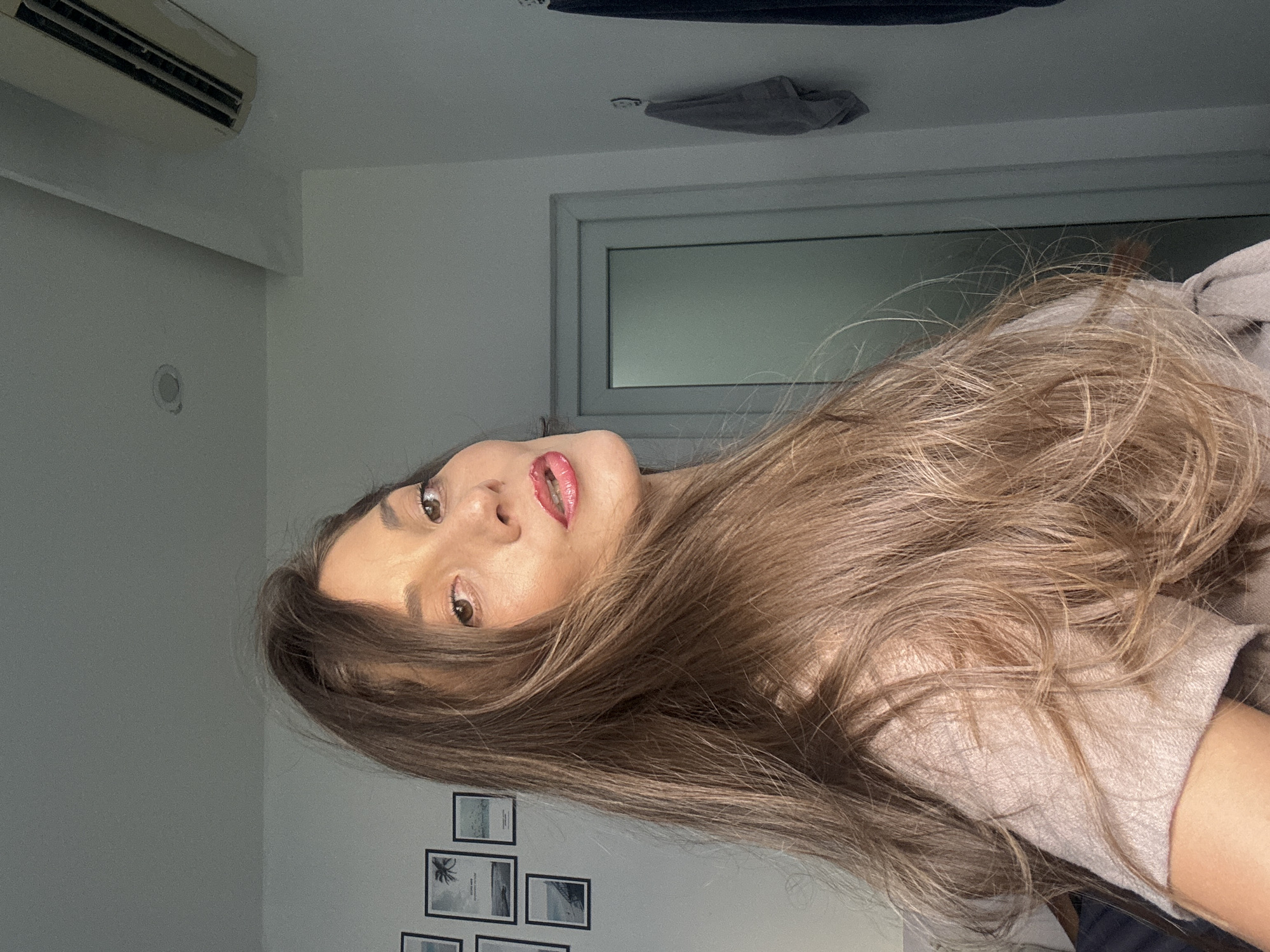 A woman with light brown hair, pink lipstick, and fair skin is positioned in the foreground, her face angled towards the camera. She appears to be indoors. Behind her are elements of a domestic interior including a white wall, a set of framed pictures depicting seaside scenes, a window, and an air conditioning unit. The location is not identifiable as Đà Nẵng, Vietnam from these elements alone. There are no actions, interactions, or other subjects apparent in the frame.