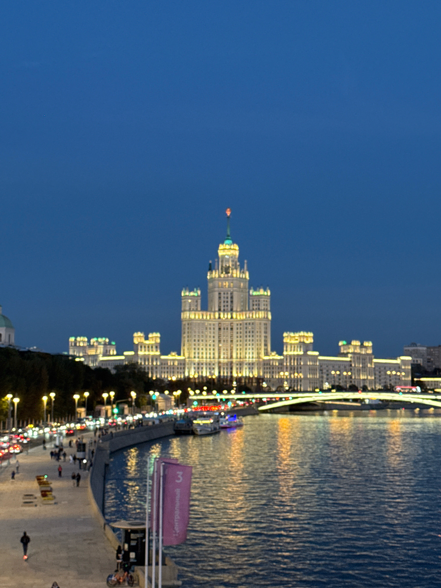 This is a nighttime photograph of the Kotelnicheskaya Embankment Building in Moscow, Russia, viewed from across the Moskva River. The building, a prominent Stalinist skyscraper, is illuminated with warm yellow lights, highlighting its intricate architecture. The Moskva River in the foreground reflects the city lights, creating a shimmering effect on the water. Along the embankment, a promenade is visible, lined with trees and streetlights, and a few people can be seen walking. Cars are driving on a road to the left of the embankment, their headlights creating streaks of light. A modern bridge arches over the river in the middle ground, also illuminated. The sky is a deep, clear blue, indicating dusk or early night.