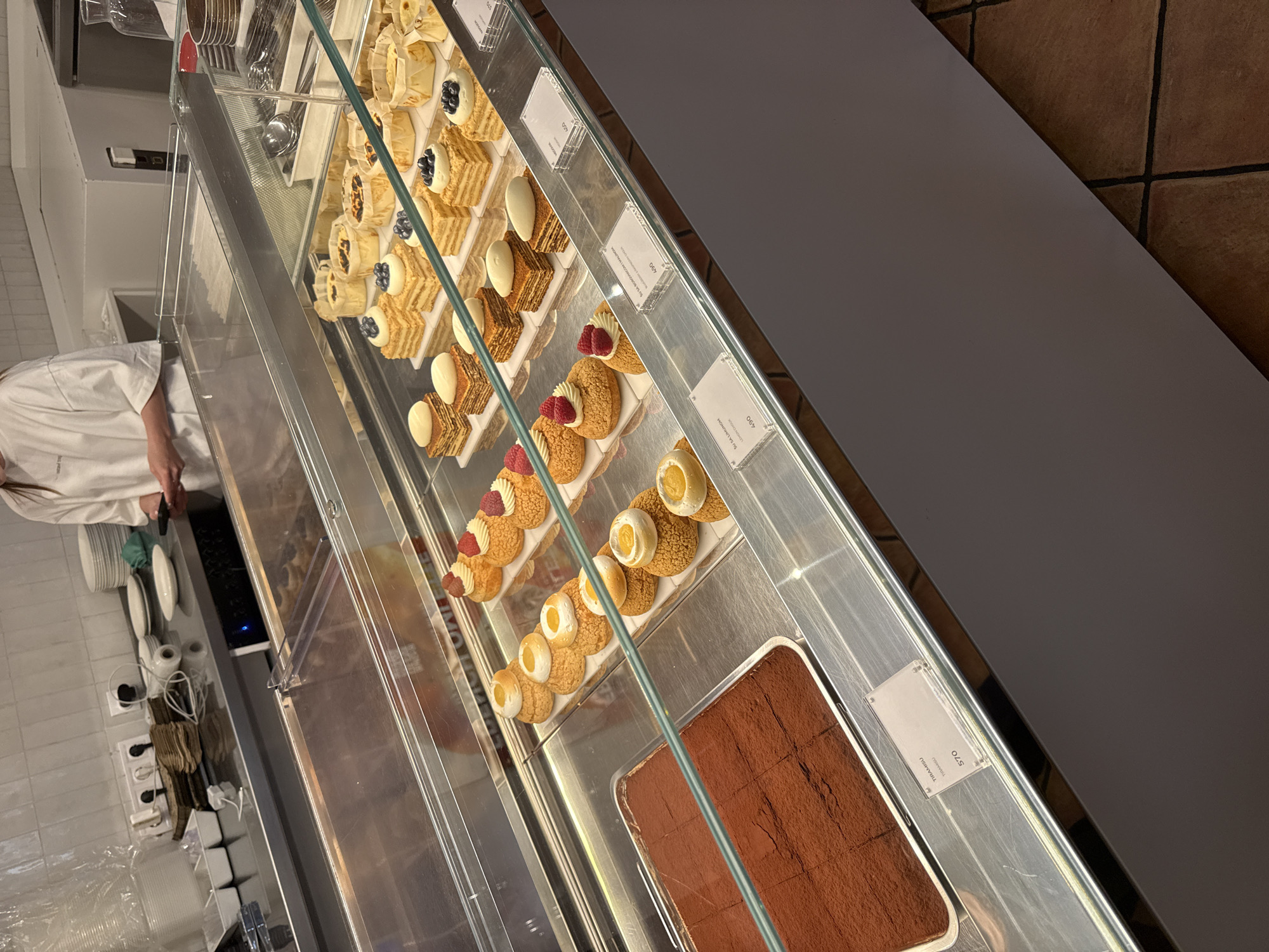 The image shows a display case filled with various pastries in what appears to be a bakery or cafe. In the foreground, a tray of rectangular chocolate cakes dusted with cocoa powder is visible. Behind this, several rows of individual pastries are arranged. These include small square cakes topped with cream and fruit, choux pastry buns filled with cream and topped with fruit or a yellow dollop, and what appear to be layered cakes with cream and berries. Several small signs with names and prices are placed in front of the pastries. In the background, a person wearing a white t-shirt is visible, holding a smartphone and standing behind a counter. On the counter, there are stacks of plates, electrical outlets, and other kitchen items. The overall environment suggests a commercial food establishment. The specific location in Belgrade, Serbia cannot be determined from the image alone.