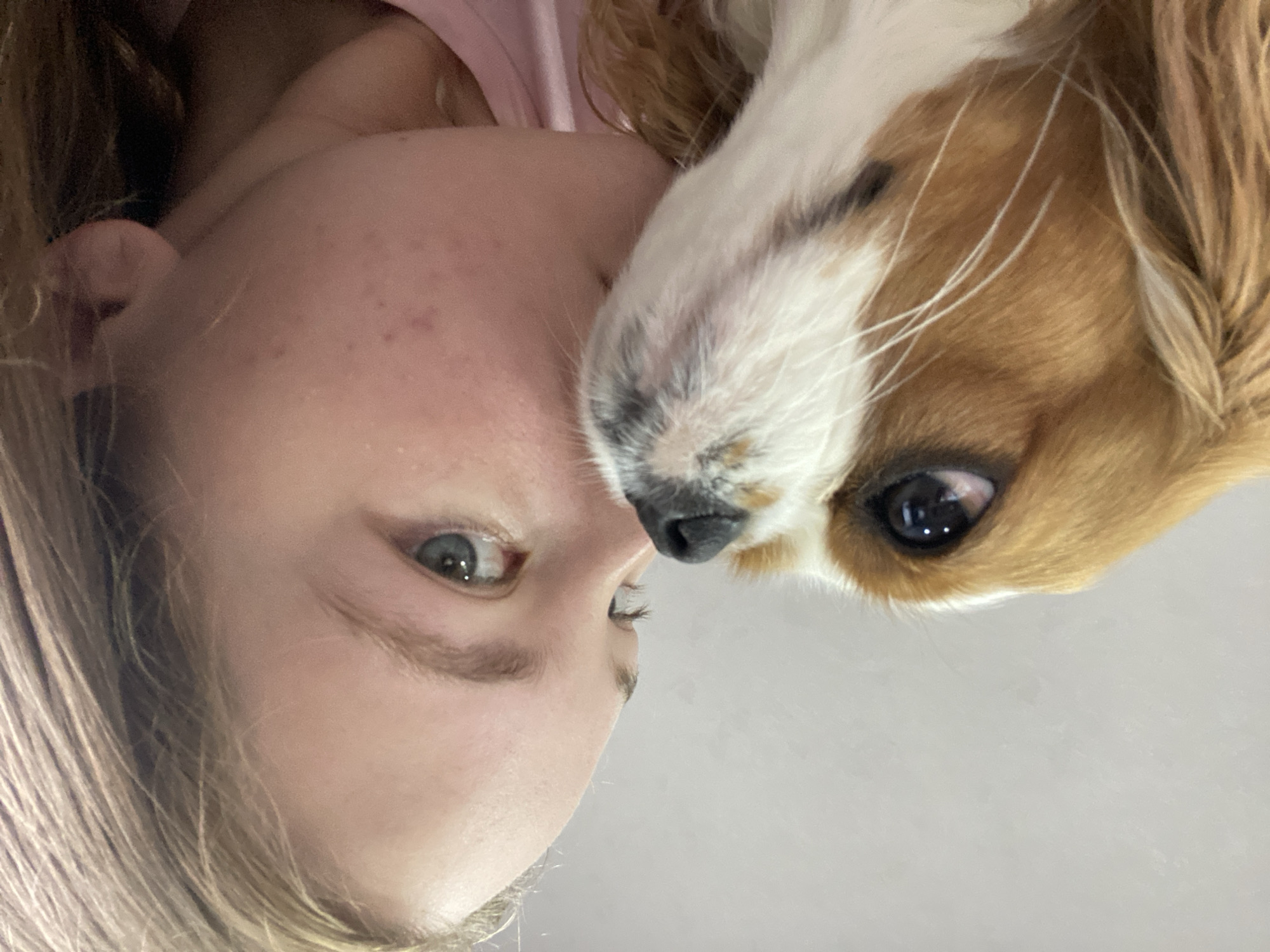 An extreme close-up shot shows a young person and a dog. The person's face is on the right side of the frame, with their eyes looking to the left. Their skin has some blemishes. The dog's face is on the left side of the frame, with its nose and eyes visible. The dog appears to be nuzzling the person. The background is a plain, light gray wall. The overall lighting is soft and even.