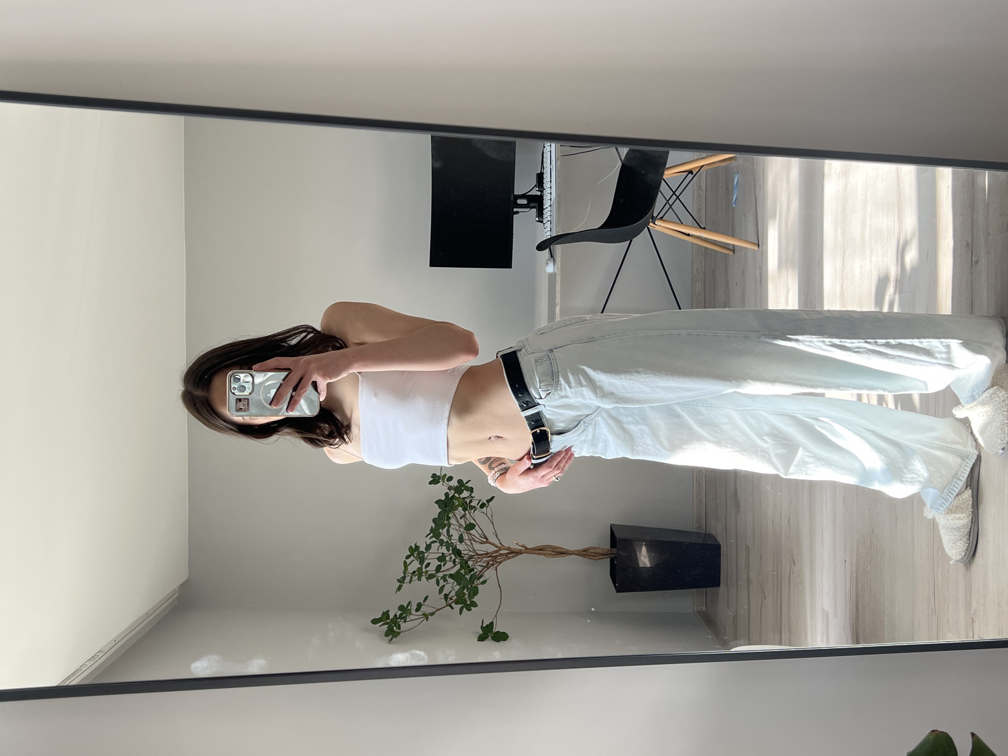 In a room with light-colored walls and a wooden floor, a young woman stands in front of a full-length mirror, taking a selfie with her phone. She is wearing a white tube top, light blue wide-leg jeans, a black belt, and fluffy slippers. Her dark hair is shoulder-length, and she has a tattoo on her hip and stomach. In the reflection, a computer monitor and chair are visible, as well as a tall plant in a black pot. The room appears to be a domestic setting, possibly an apartment in Ternopil, Ukraine, given the context of the request. The overall scene is casual and relaxed, illuminated by natural light streaming in from a window.