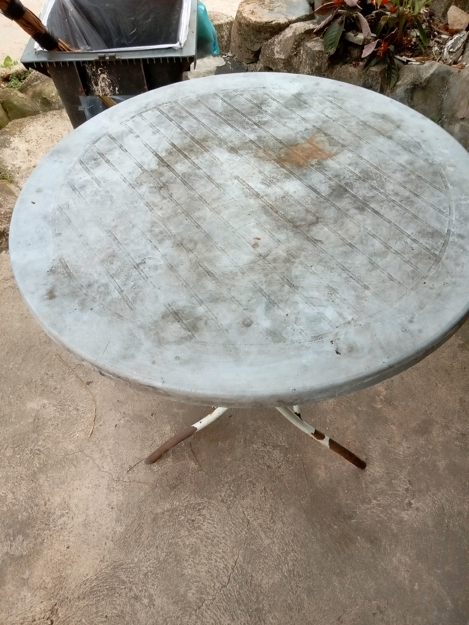 An outdoor, slightly elevated, round, gray plastic table with a criss-cross textured surface rests on a concrete ground. The table has a weathered appearance with visible scuff marks and dirt. Its white metal legs, some of which appear rusted at the ends, are splayed outwards. To the left of the table, a black plastic trash can with a lid is visible. A broom is partially inserted into the trash can. In the background, rocks and some green foliage are present. The scene is likely an outdoor residential or public area in Batam, Indonesia, given the style of the table and the surrounding environment.
