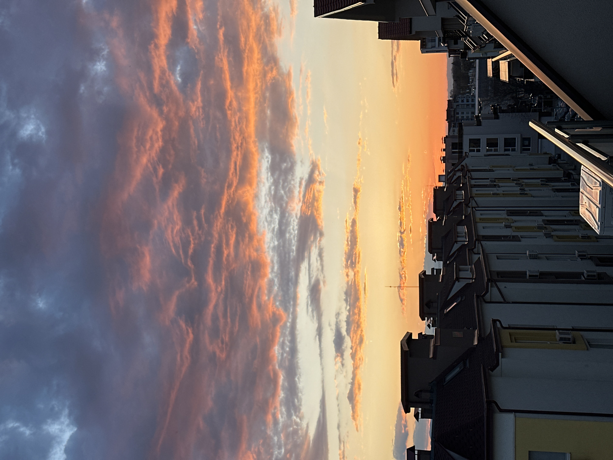 The image captures a sunset scene in Plovdiv, Bulgaria, with residential buildings in the foreground. The sky is dominated by colorful clouds, transitioning from darker, potentially stormy clouds on the left to bright orange and yellow hues near the horizon on the right. The buildings are mostly white or light-colored with dark roofs, and appear to be multi-story apartment complexes. There is a tall antenna visible near the center of the frame, possibly a TV or radio transmission tower. The buildings line the horizon with windows showing on the left side, suggesting people are inside. A balcony in the extreme upper right corner shows a portion of railing, the edges of the picture show shadows of buildings and overhangings.