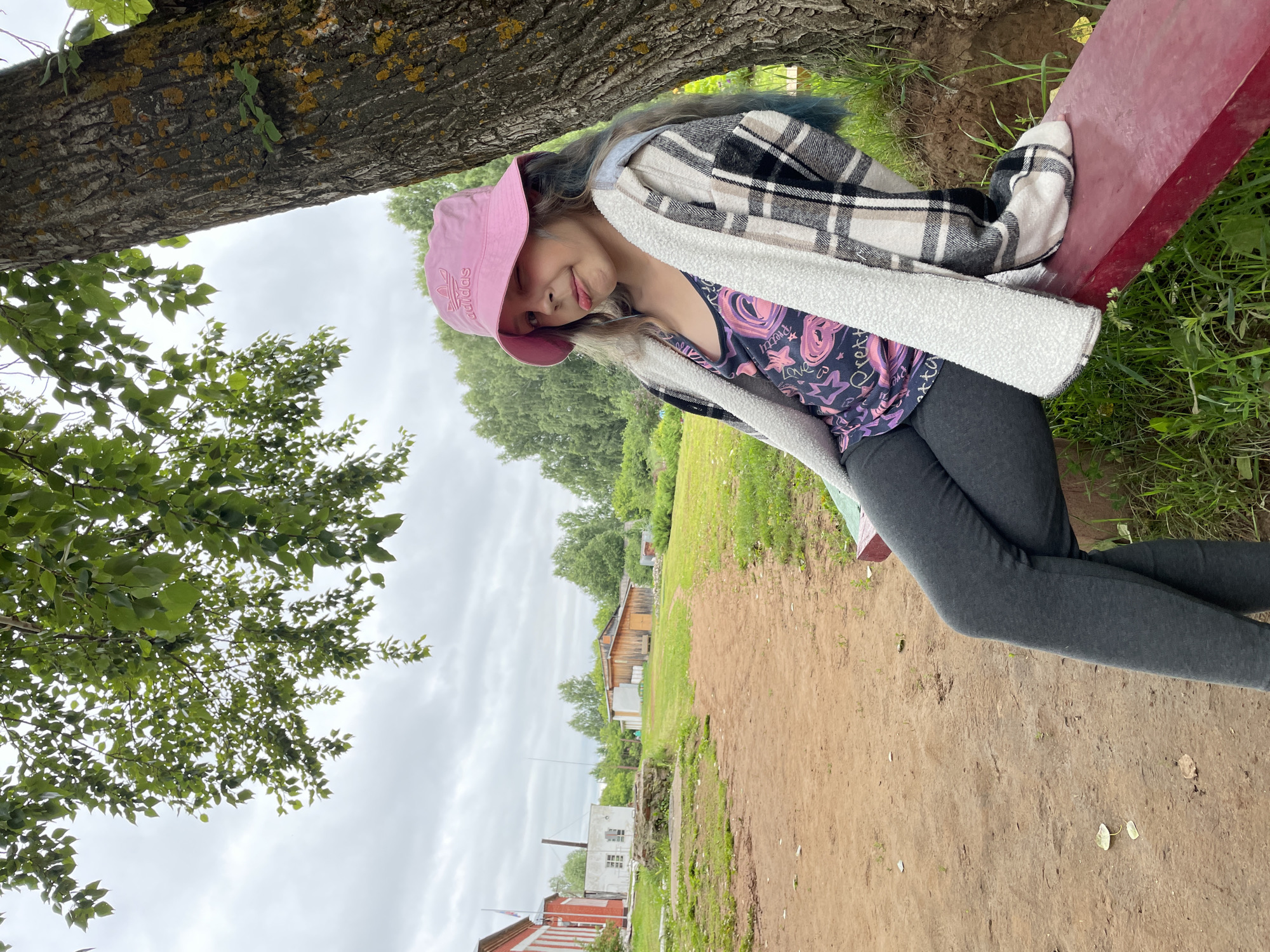 In the village of Baldeika, Russia, a young girl wearing a pink bucket hat, a patterned tank top, a plaid jacket, and dark leggings sits on a red bench next to a large tree. Her left leg is crossed over her right, and she is sticking out her tongue, looking towards the camera. The background consists of a dirt path, green grass, trees, and several small buildings. The sky is overcast.