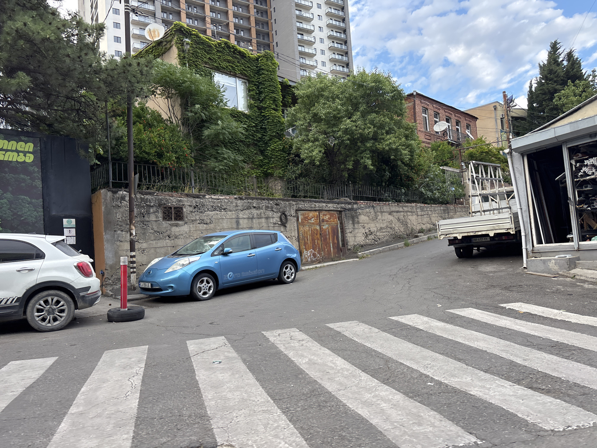 This media file depicts a street scene in Tbilisi, Georgia, under a cloudy sky. In the foreground, a pedestrian crosswalk is visible, with its white stripes painted on the asphalt. To the left of the crosswalk, a white SUV is parked, partially obscuring a black vertical sign with green text in an unfamiliar script.
Further into the scene, a blue electric car is parked near a red and white traffic bollard that sits atop a spare tire. The car has "Zero Emission" written on its side. Behind the cars, a weathered concrete wall runs along the street, topped by a metal fence. Beyond the fence, a building covered in ivy stands out, with a modern multi-story building rising behind it.
To the right, a white truck with a flatbed is parked, its license plate visible as "HH 417-VV". Adjacent to the truck is a commercial building with large windows, appearing to be a shop. The overall impression is of a somewhat urban and slightly aged streetscape, common in many cities. The time of day appears to be daytime, given the natural light, and the weather is overcast. No people are visible in the frame.