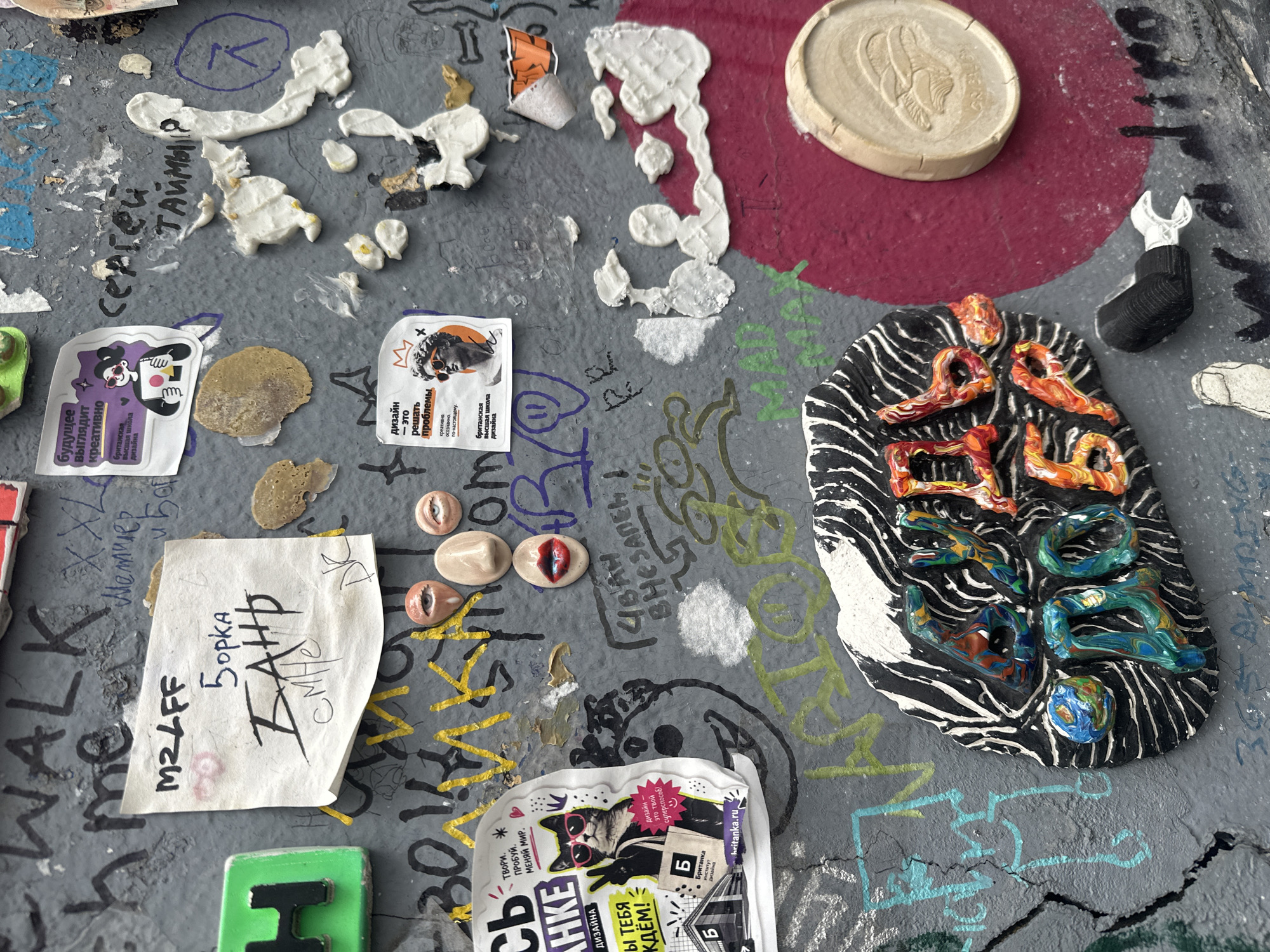 This is a close-up shot of a textured, possibly concrete, surface, likely a wall or sidewalk, heavily adorned with affixed objects, stickers, and graffiti. There are various stickers with Russian text. One sticker featuring a cat in sunglasses with the phrase "Дизайн - это твой суперспособ!" (Design is your superpower!). Another sticker says "Будущее выглядит креативно." (The future looks creative). A piece of paper with handwritten text including "M2LFF" and "Борка". Several ceramic elements are attached to the surface, including a round disc, a set of sculpted eyes, and a black and white striped piece with colorful swirl shapes. There is also a miniature wrench emerging from a sock. The Russian phrase "4BAH BHEЗAПEH" (Ivan Suddenly) is scrawled on the surface. The location may be in Moscow, Russia, given the Russian text present on the stickers and handwritten notes.