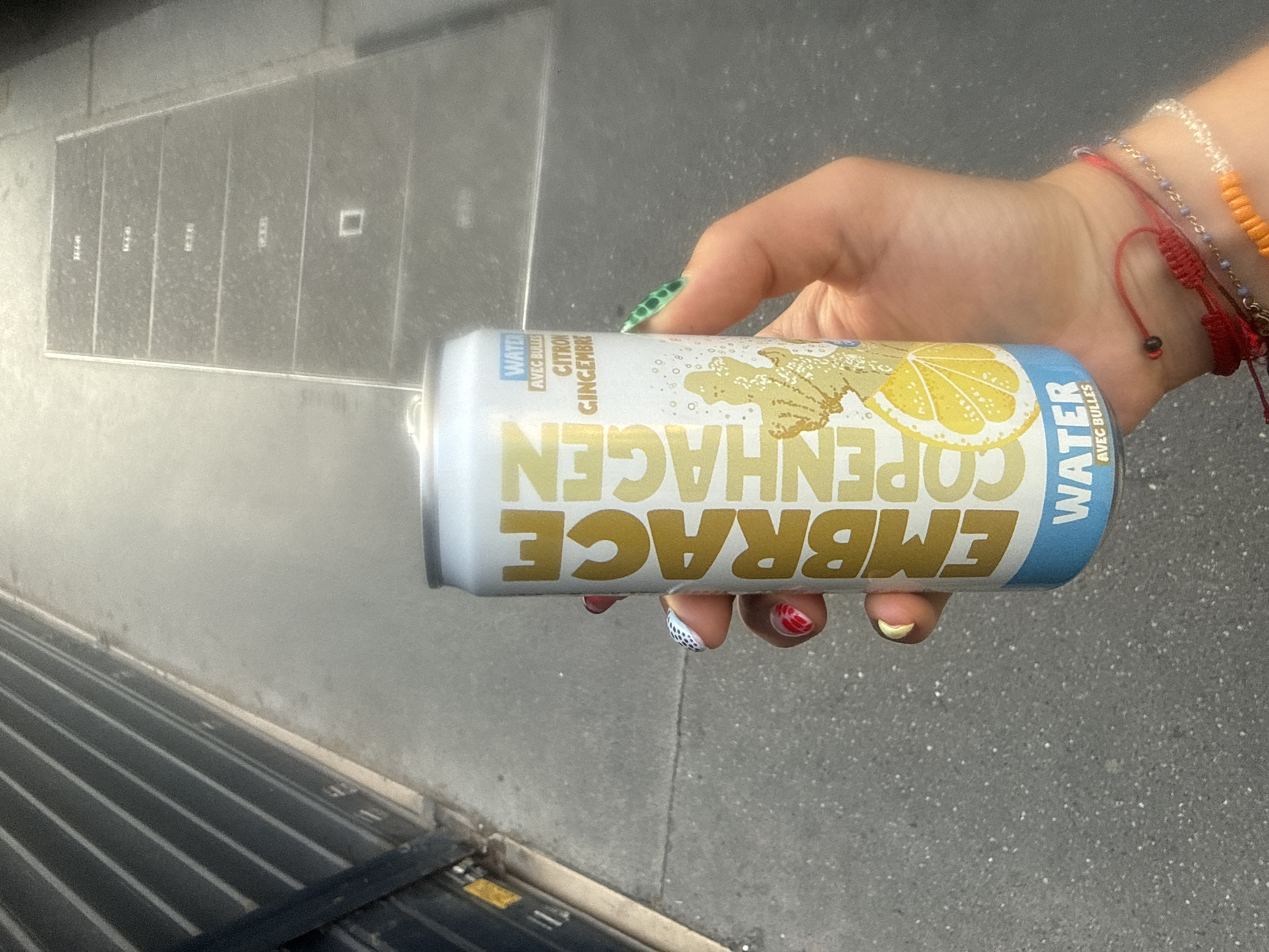 A person's hand with painted fingernails holds a can of "EMBRACE COPENHAGEN WATER CITRON GINGEMBRE" (Lemon Ginger) outdoors. The can is white with gold lettering and a blue band at the bottom. The background shows a textured gray concrete surface, possibly a sidewalk or street, with a dark metal structure on the left and a manhole cover or utility panel with linear divisions on the right. The lighting suggests it is daytime, with shadows indicating a sun position. The presence of a manhole cover and general urban infrastructure suggests this scene is located in a city, consistent with a Parisian street.