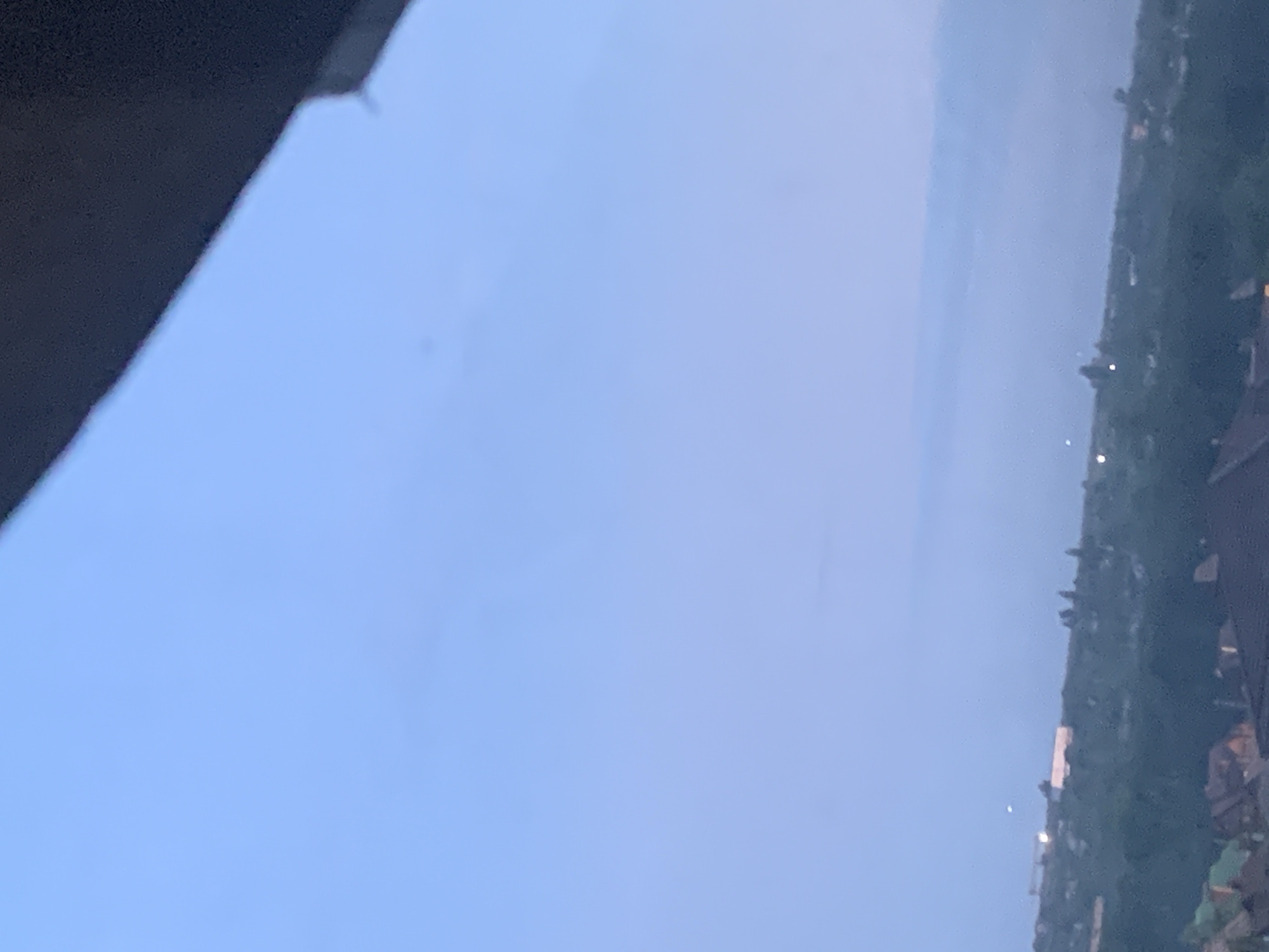 A wide shot depicts a hazy blue sky with a dark overhang in the upper right corner. The horizon line is low in the frame, showing a blurry expanse of green trees and scattered buildings. The scene appears to be an outdoor view from an elevated position, possibly a balcony or rooftop. Lights are visible in the distance, suggesting the presence of a settlement. The overall atmosphere is serene and slightly overcast, with a hint of twilight.