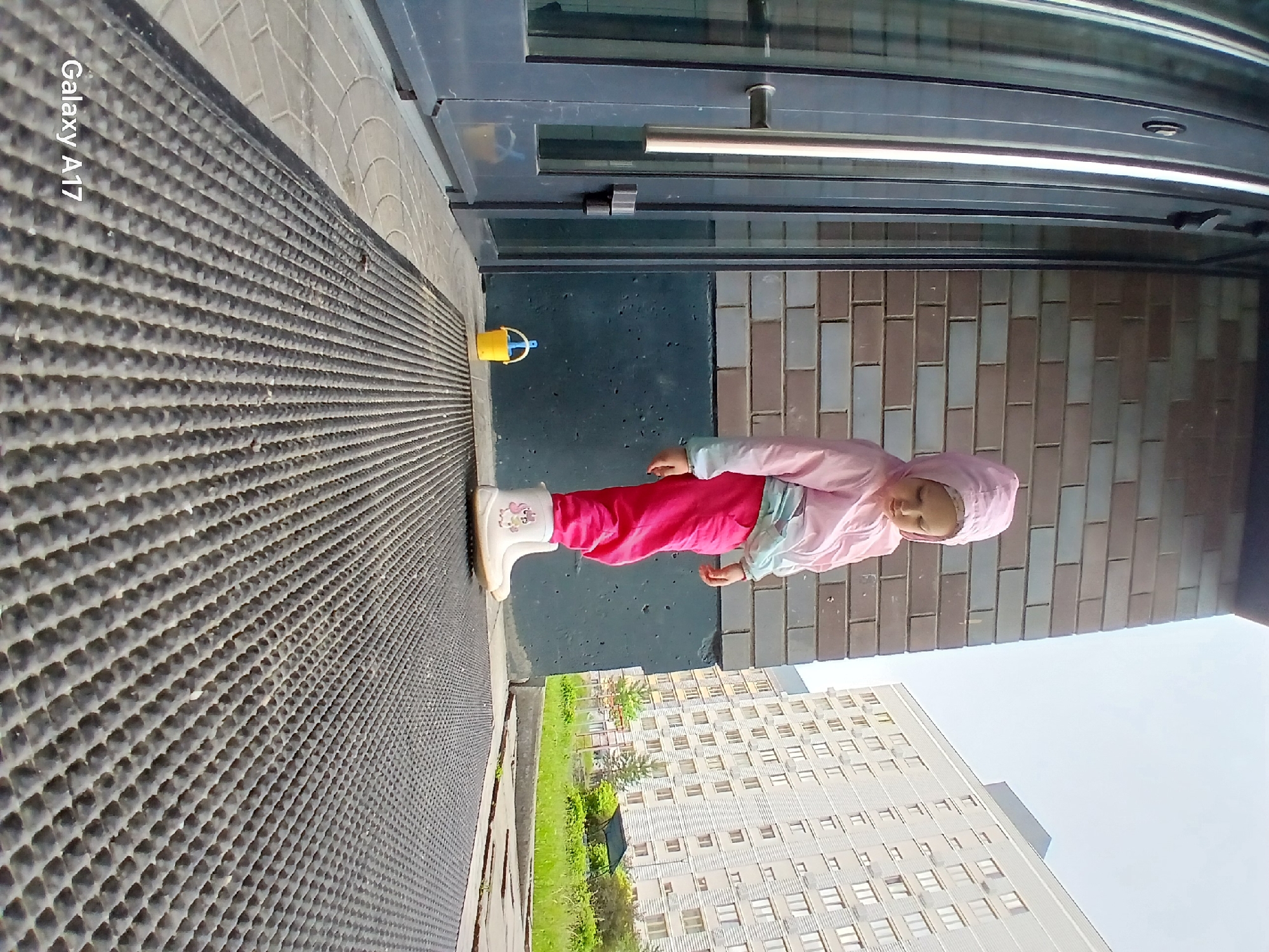 An outdoor scene in an urban residential environment, consistent with a district in Yekaterinburg, Russia. A young child, appearing to be a toddler or preschooler, stands centered in the mid-ground. The child wears a light pink hooded jacket with a white-to-light-blue gradient section, bright pink trousers, and white rubber boots decorated with a pink unicorn and rainbow motif on the left boot. The child is facing slightly left, looking downwards.
To the child's left is a dark-framed glass entrance door with a prominent silver horizontal handle and a black lock mechanism. The door reflects the surroundings. Adjacent to the door, a brick wall rises, composed of alternating light and dark brown/grey bricks. Below the brickwork is a dark blue-grey painted or textured wall section.
A small, bright yellow plastic bucket with a light blue handle and a matching small blue scoop/shovel inside rests on the paved ground between the door and the child. The foreground is dominated by a large, dark grey rubber mat with a uniform pattern of raised circular nubs. To the left of the mat, the ground is paved with light-colored tiles featuring a circular design.
In the background, beyond a strip of green grass and scattered trees/bushes, multiple large, light-colored, multi-story residential buildings with numerous windows are visible under an overcast sky. A dark, possibly covered structure is partially visible within the green area. The image includes a "Galaxy A17" watermark in the bottom left corner.