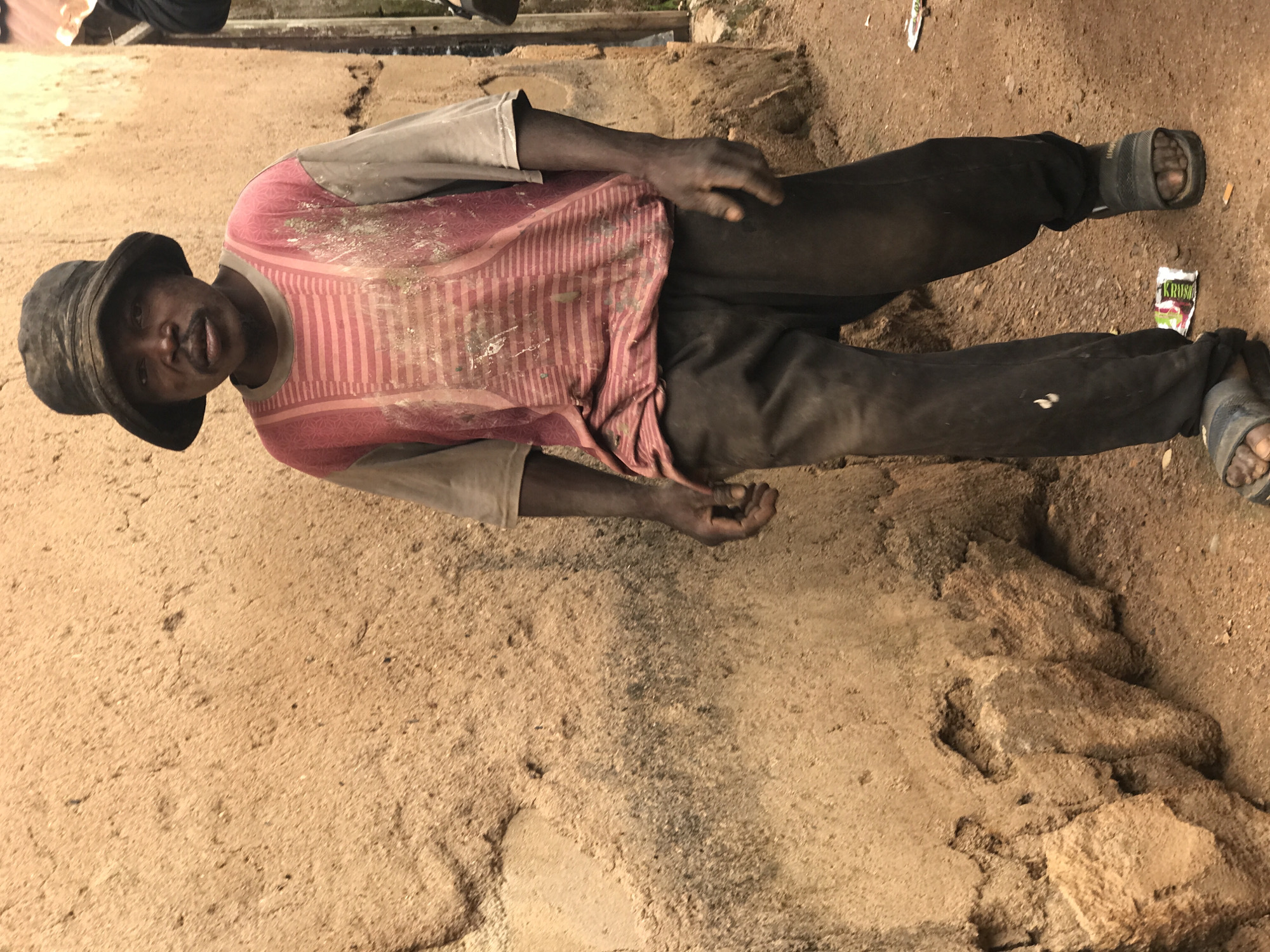 A medium shot captures a man wearing a dark bucket hat and a dirty pink and beige t-shirt. He stands in front of a rough, textured, beige wall that appears to be made of mud or clay. The man is looking directly at the camera. His t-shirt has horizontal stripes in shades of pink and lighter pink, with some visible dirt or stains. His pants are dark brown and appear worn and dirty. He is wearing dark sandals on his feet. The ground in front of him is a sandy, dirt path with some small debris, including a crumpled packet with writing on it. To the right of the man, a portion of a dark, vertical object, possibly a pole or part of a structure, is visible. The overall environment suggests an outdoor setting in a possibly rural or underdeveloped area.