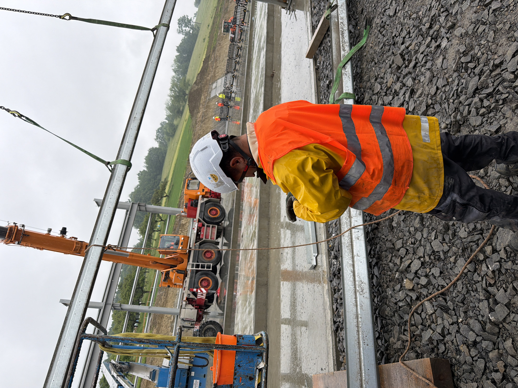 A construction worker wearing a white hard hat, a yellow waterproof jacket, and an orange high-visibility vest stands on a gravel surface, looking down at a handheld device. The worker is in the foreground, facing away from the camera. In the background, an orange crane is lifting a large steel beam. Other construction equipment, including a cherry picker and a truck, are visible. The scene appears to be a construction site, possibly in Janowice, Poland, with overcast skies and wet ground, indicating recent rain. The environment suggests ongoing construction of a large structure, likely industrial or infrastructure-related, given the scale of the materials and equipment.