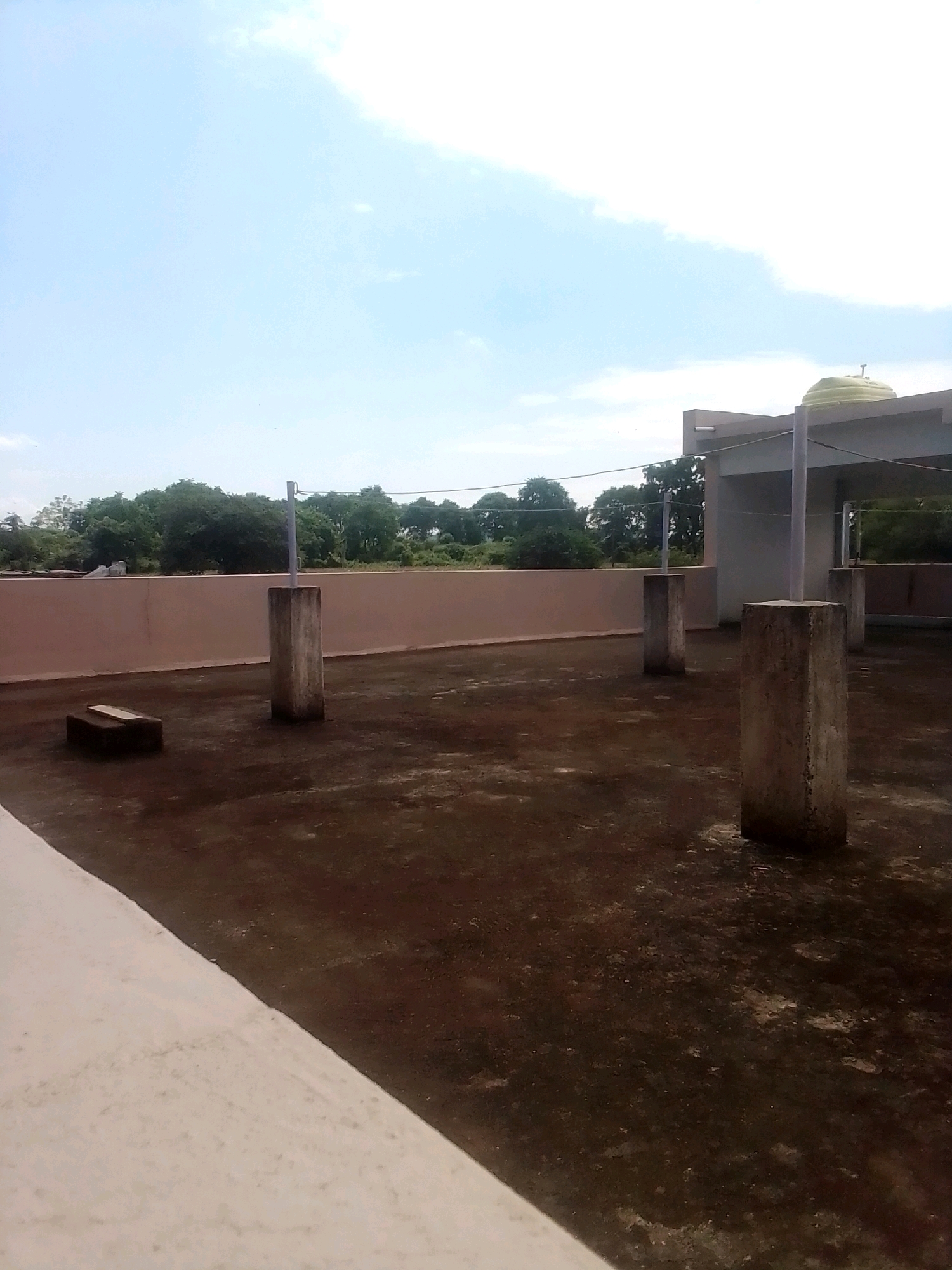 The image depicts a rooftop in Anjaniya, India, under a bright, partly cloudy sky. The rooftop is paved with a dark, mottled material and features several concrete pillars of varying heights. In the foreground, a white, angled parapet wall is visible on the left. To the left of the frame, on the rooftop surface, is a dark rectangular object with a light-colored plank placed on top. A pinkish-brown wall with trees and vegetation visible behind it forms the background. In the distance, a light-colored building with a dome-shaped water tank on its roof is present. The scene appears to be quiet and undisturbed, with no people or animals present.