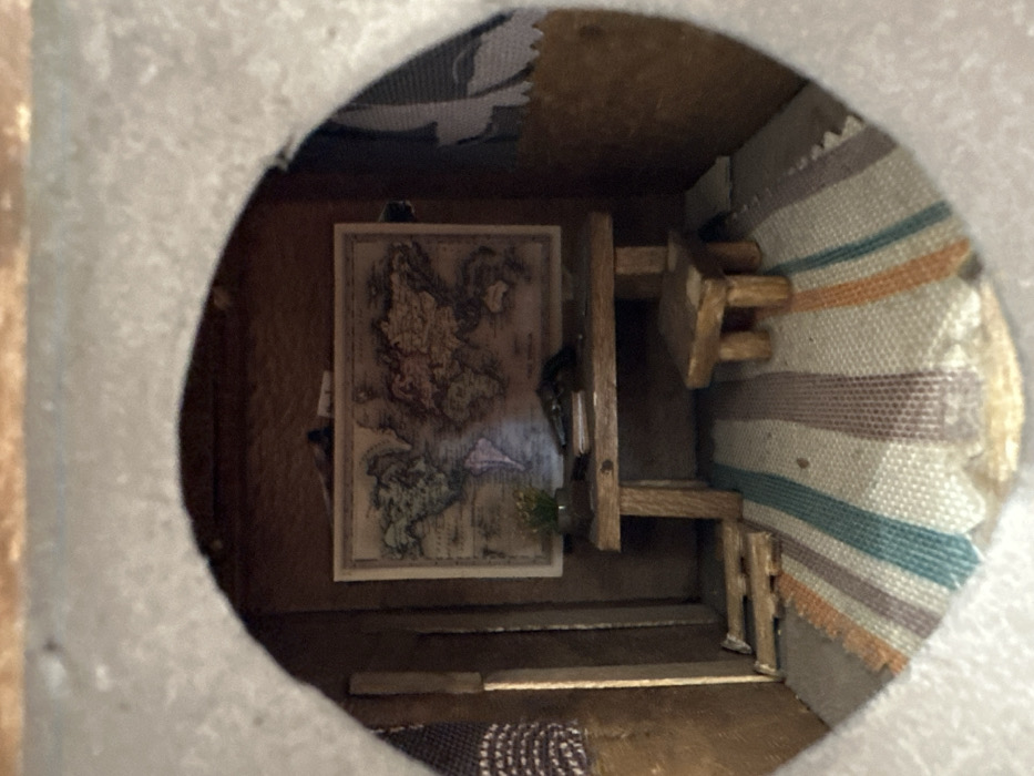 The image captures a detailed view inside a miniature room or diorama, observed through an irregular, semi-circular opening in a light-colored material. The overall impression is that of a small, enclosed space, possibly a dollhouse interior.
On the back wall, a framed, rectangular picture of an antique-style map is prominently displayed. The map features intricate landmasses and details, rendered in earthy tones of brown, green, and beige, suggesting either a historical map or a fantasy realm. To the right of the map, a small, multi-tiered wooden structure, possibly a shelf unit or part of a miniature furniture piece, stands against the wall. Further to the right, the wall is adorned with a striped fabric, resembling a miniature rug or tapestry, showcasing horizontal bands in colors such as beige, light blue, orange, and brown. The floor of the room appears to be made of wood, and a small segment of a patterned rug or mat with brown and lighter tones is visible in the foreground. A tiny green plant-like object is situated near the bottom center, in front of the map.
No people are visible, and there is no explicit activity taking place within this static scene. The lighting suggests an external source illuminating the interior, casting some light on the map and surrounding objects. No legible text is discernible on the map or elsewhere in the image. The setting conveys a sense of a carefully crafted miniature environment. The photograph was taken in Metamorfosi, Greece.