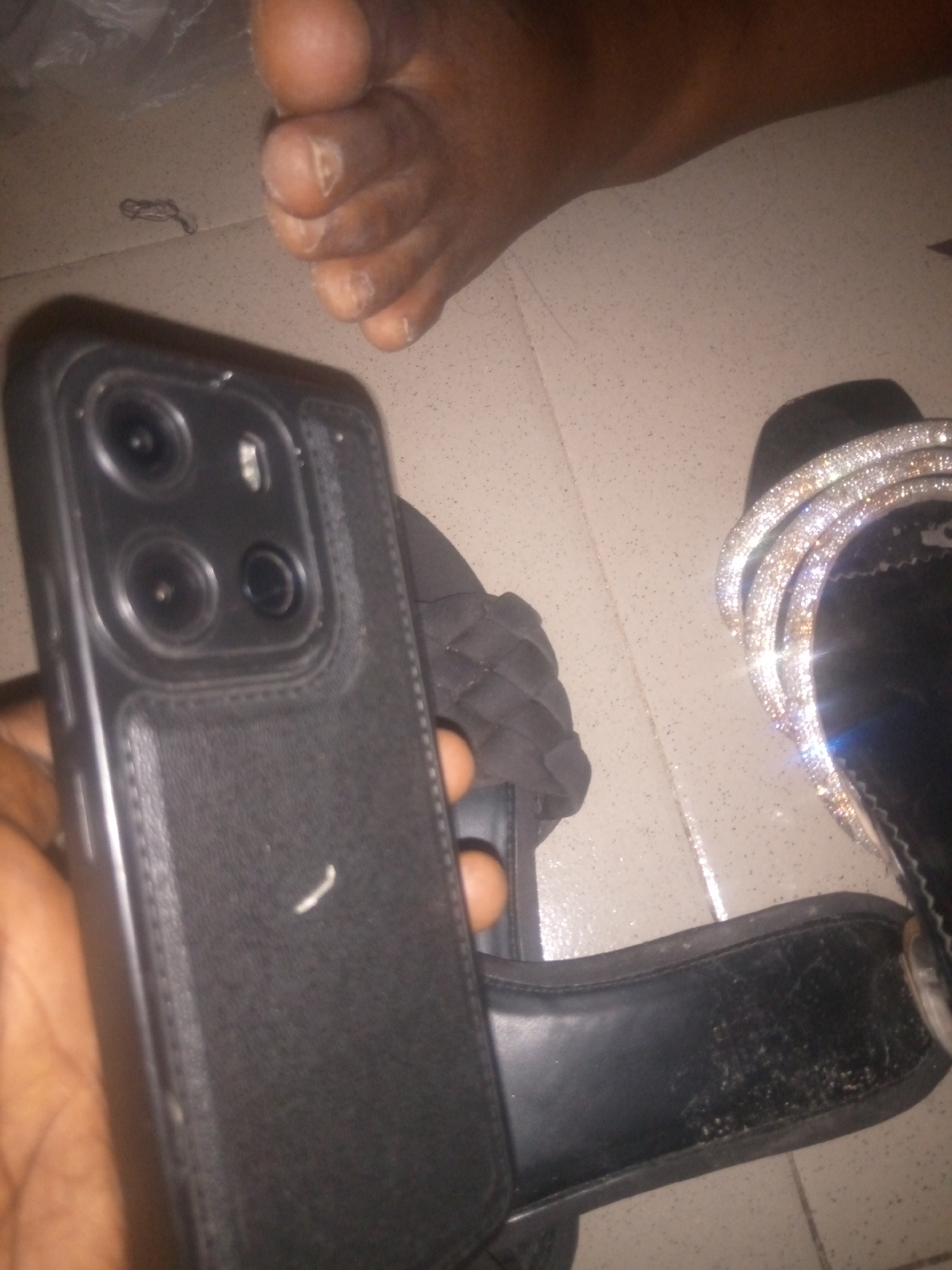 In Jauro Yinu, Nigeria, a person's foot, with visible toenails, rests on a tiled floor. Next to it, a black iPhone 14 Pro Max, held by an unseen hand, is angled upwards, revealing its camera module. A black sandal adorned with rhinestones is placed to the right of the phone, and another black sandal with a textured strap lies to the left of the phone. The environment appears to be indoors, possibly a home or a shop.