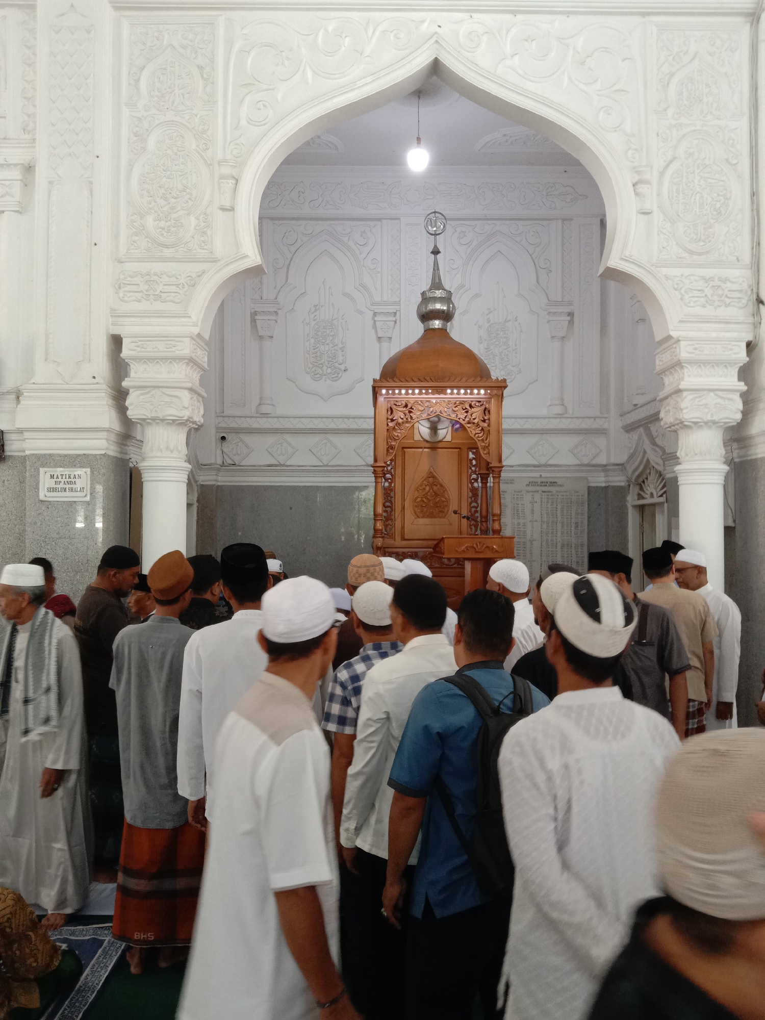 Inside a mosque in Lhokseumawe, Indonesia, a group of men, some wearing skullcaps, are gathered. The interior architecture is characterized by white archways and pillars with ornate carvings. A wooden pulpit is centered in the scene, beneath a light fixture and a religious symbol. A sign is visible on a column, and a list is displayed on the wall behind the pulpit. The men are dressed in various styles, including traditional robes, shirts, and trousers. Some are facing forward, while others are looking around or interacting. The floor is covered with patterned carpets.