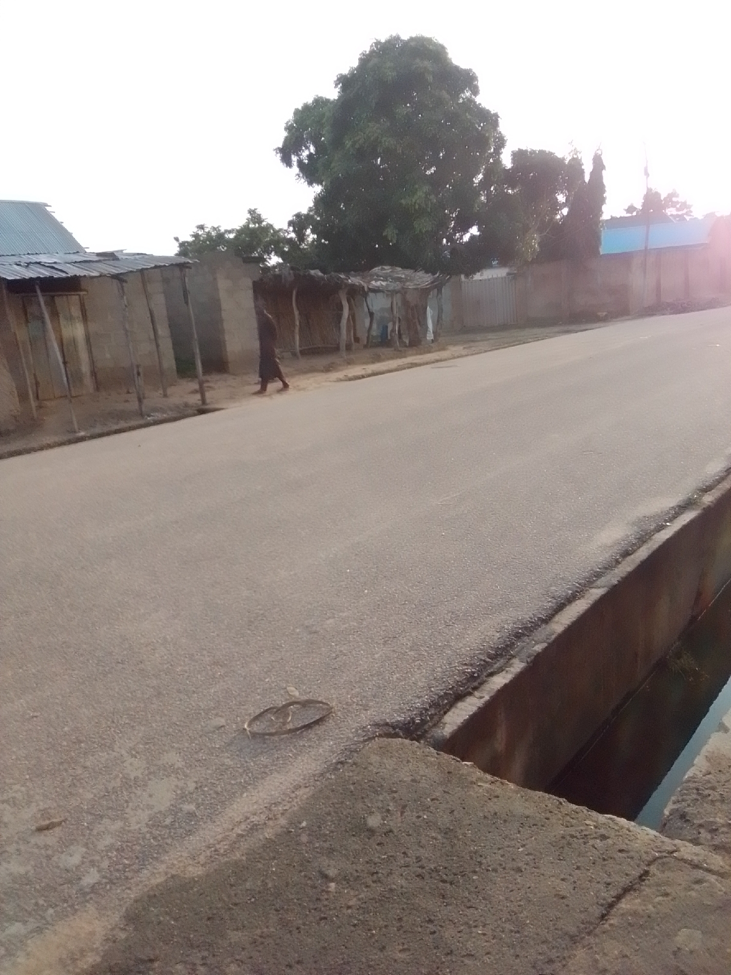This image captures a street scene in Gombe, Nigeria. A person dressed in dark clothing walks along the side of a paved road. To the left of the road are several structures with corrugated metal roofs and mud or brick walls, some appearing to be makeshift dwellings or small shops. Beyond these structures, large trees and foliage are visible. Further in the background, a building with a blue roof and a wall can be seen. The right side of the road features a concrete drainage channel, partially filled with water. The overall environment suggests a semi-urban or rural setting within the city.