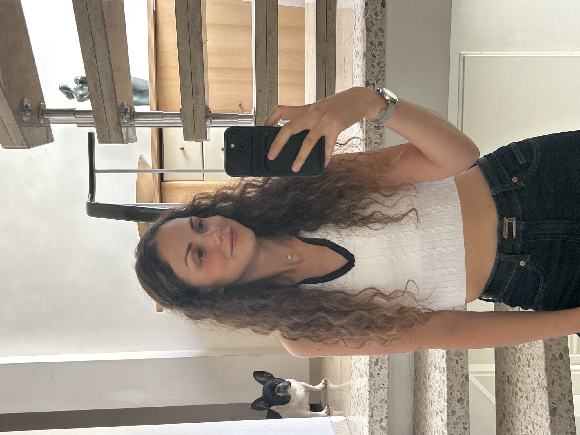 A young woman with long, curly brown hair takes a mirror selfie in a well-lit indoor space. She is wearing a white crop top with a black trim around the neckline and dark wash jeans. On her left wrist, she wears a silver watch. She holds a black smartphone in her right hand to capture the image. Behind her, a staircase with metal railings and stone steps is visible. To the left of the staircase, a black and white French bulldog stands on a lower step, looking towards the camera. The background includes a wall with a white pillar and wooden cabinets.
