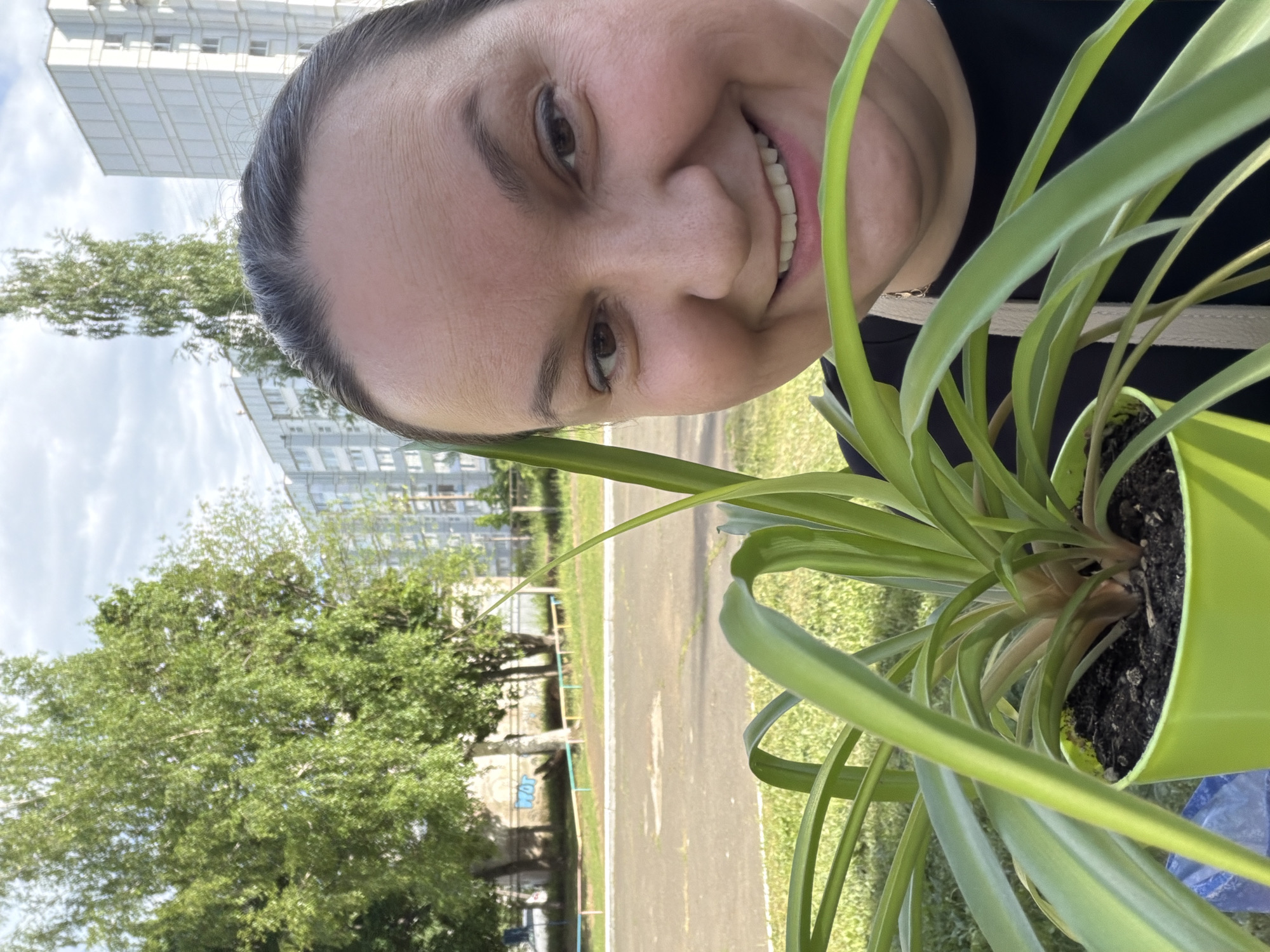 A smiling woman with fair skin and dark hair is holding a potted plant with long, green leaves. The pot is bright green and filled with dark soil. Behind her, there is an outdoor scene with green trees, grass, and a building in the background, possibly apartments or a residential building. A road or path is visible in the mid-ground. The photo was taken in the city of Chaikovsky, Russia.
