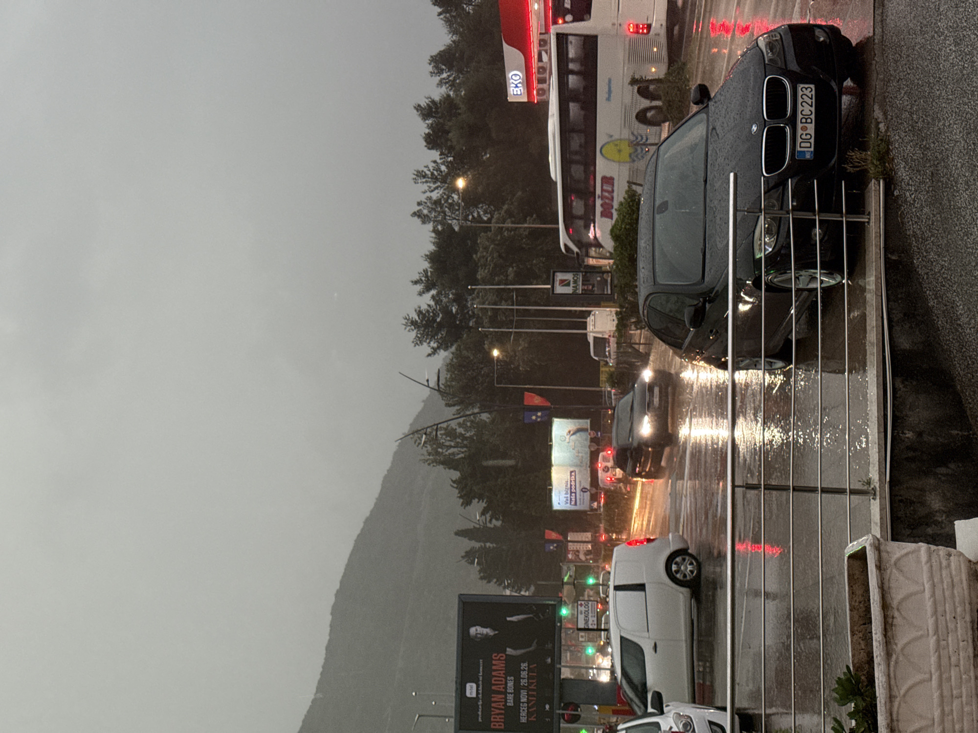 The image captures a rainy street scene in Budva, Montenegro, during what appears to be an overcast day or early evening, characterized by a grey sky and wet, reflective roads.
In the foreground, a dark-colored BMW sedan is parked or stopped beside a metal barrier, facing away from the viewer towards the left (when the image is viewed upright). Water droplets are visible on its surface, indicating recent or ongoing rain. Its license plate reads "DG BC223", confirming a Montenegrin registration (DG for Podgorica, likely as Budva often uses DG plates due to proximity or specific registration practices). Next to the barrier, a concrete planter is visible.
To the left of the BMW, a busy street is visible with vehicles and illuminated signs. A white bus with red and white livery is prominent, featuring the text "BOŽUR" and "MTB" on its side, and "EKO" on a red sign above it. Other cars, including a white van, are visible further down the road, their headlights and taillights creating reflections on the wet asphalt. Traffic lights are illuminated, showing green.
Several billboards and signs line the road. One large billboard prominently features "BRYAN ADAMS BARE BONES HERCEG NOVI / 26.06.26. KANLIKULA," advertising a concert. Another sign displays "Val biznis. Naša podrška.," and nearby, "GINEKOLOG" and "NAMOS" are also visible. Streetlights are illuminated, casting light onto the wet road.
The background consists of a tree-lined hillside or mountain range under the same grey, overcast sky, suggesting a natural landscape surrounding the urban road. No people are explicitly visible in the frame. The overall atmosphere is one of a typical, busy road on a rainy day or evening in a town setting.