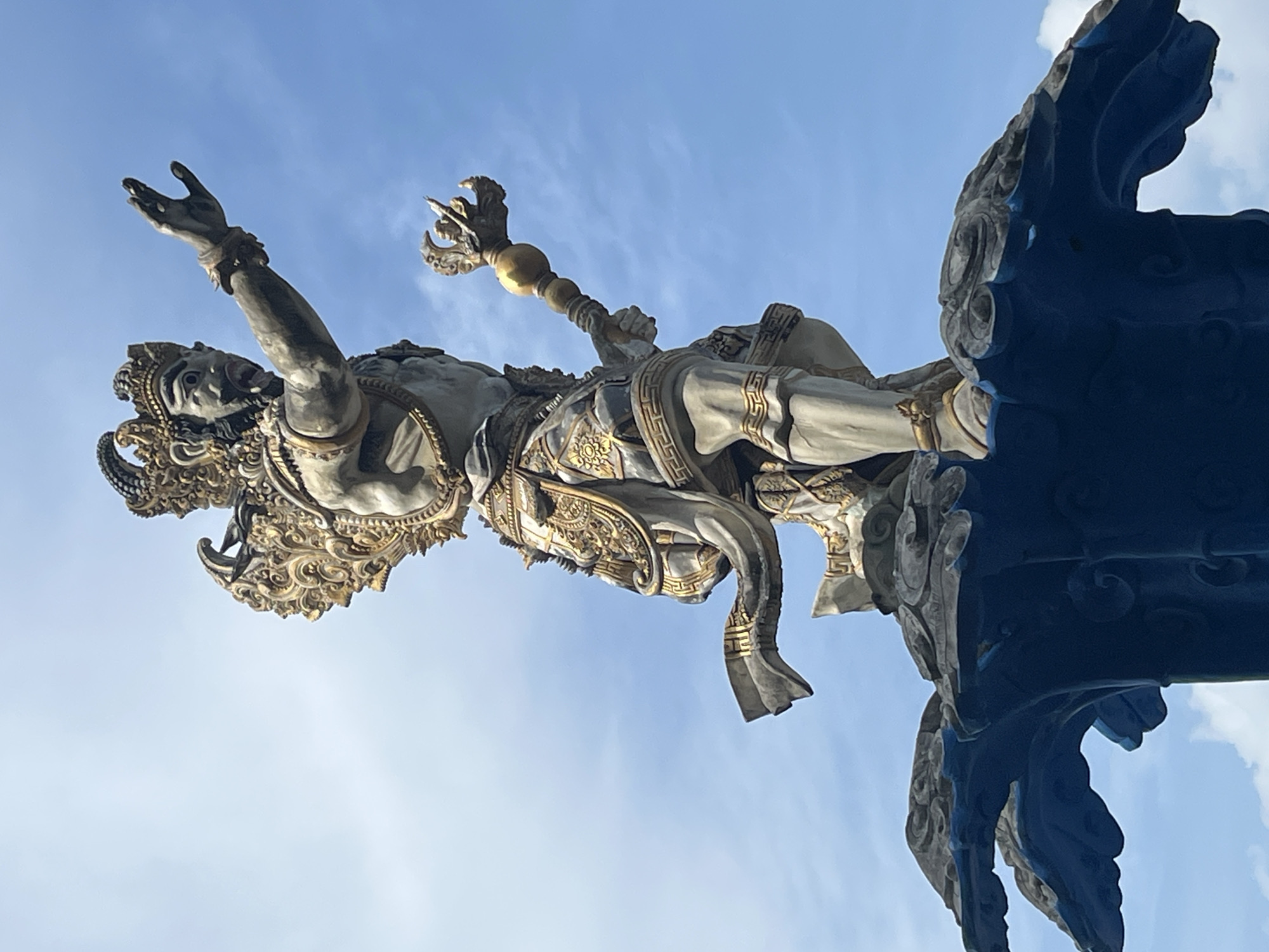 A majestic statue of a deity, possibly Garuda, stands prominently against a clear blue sky with scattered white clouds. The statue, rendered in a style suggestive of Balinese Hinduism, is depicted with a fierce expression, adorned with intricate gold detailing on its headdress, armor, and attire. It holds a golden scepter in its right hand and has its left arm extended forward. The deity is positioned atop a blue, wave-like structure with ornate carvings, which appears to be part of a fountain or monument. The overall scene captures a sense of grandeur and spiritual significance, characteristic of landmarks found in the Kuta Selatan region of Bali, Indonesia.