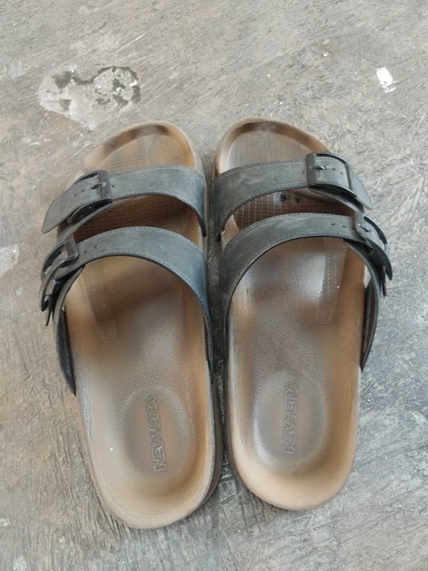 A pair of black and brown unisex sandals are resting on a concrete surface. The sandals have two adjustable straps across the foot, each with a buckle. The insoles are a light brown color and have the word "NEWMERA" embossed on them. The concrete surface is textured and appears to be outdoors, with some debris scattered around. There are no people or animals visible in the image, and no specific landmarks are identifiable. The scene does not provide any information to suggest a specific location within the city of Surabaya, Indonesia.