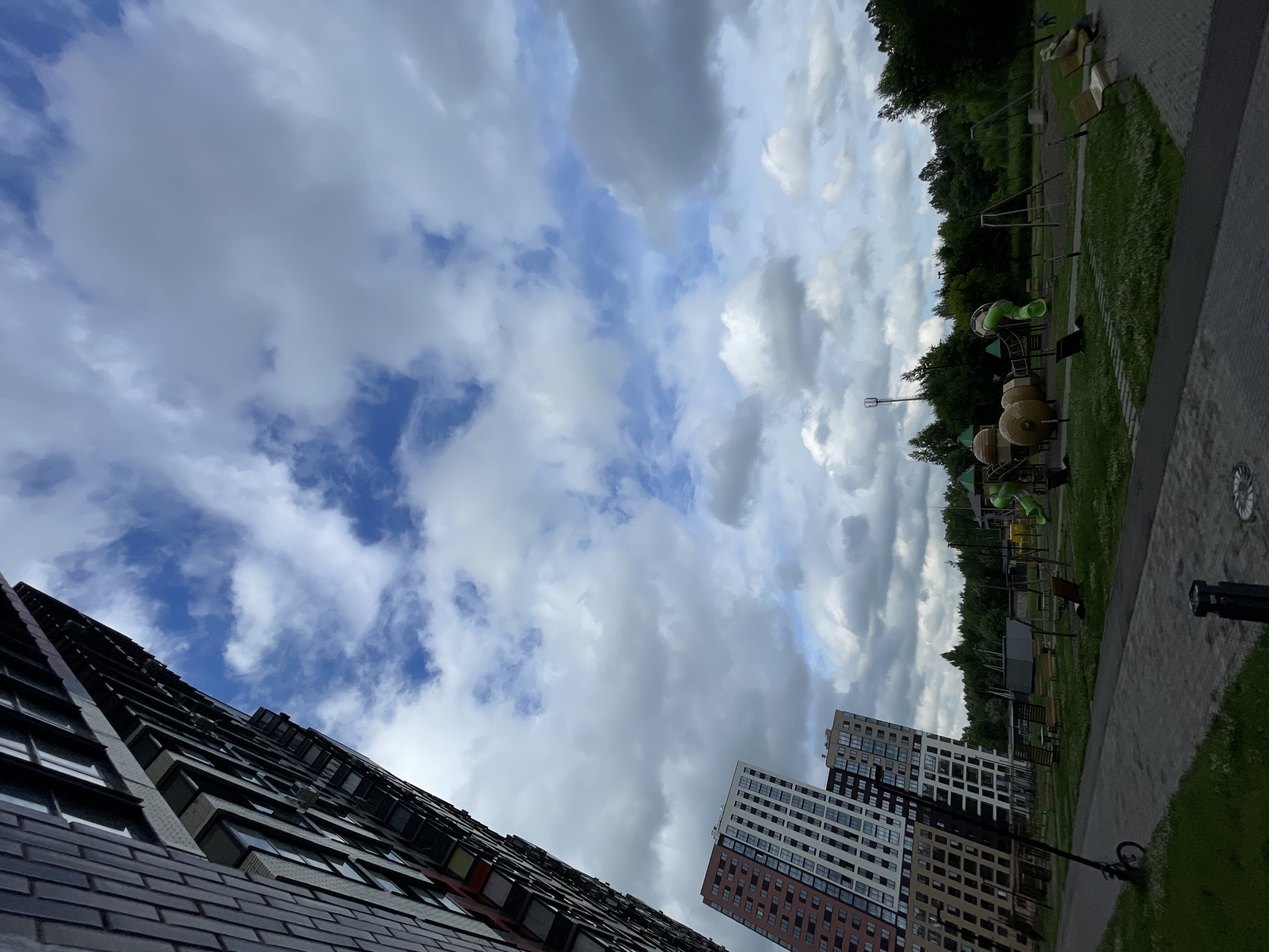 An outdoor, wide-angle, low-angle shot captures a modern apartment complex and a playground under a cloudy sky. The building on the far left is tall and dark, with visible windows and balconies, angled towards the sky. To the right of it, two slightly shorter, multi-colored apartment buildings with many windows are visible, also angled. In the foreground, a paved walkway runs alongside a grassy area with a small playground. The playground features a green slide, swings, and other play structures. Benches are scattered throughout the grassy area. Trees form a line behind the playground. A single bicycle is parked on the side of the walkway. The sky is filled with a mix of dark and light clouds, with patches of blue sky visible. The scene appears to be in a residential area.