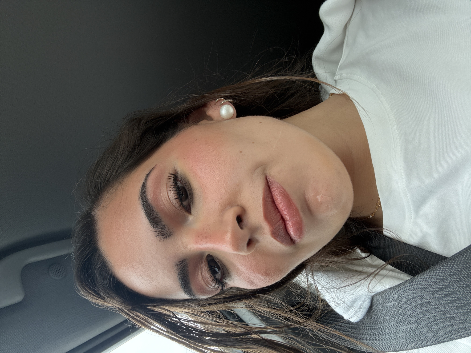 A low-angle close-up shot features a light-skinned woman with brown hair wearing a seatbelt and a white shirt. She is looking directly at the camera with her head tilted. She has dark eyebrows and eyelashes, and her lips are adorned with a pink lipstick. A single pearl earring is visible on her left ear. The background appears to be the interior of a car. The location is San Antonio, United States of America.