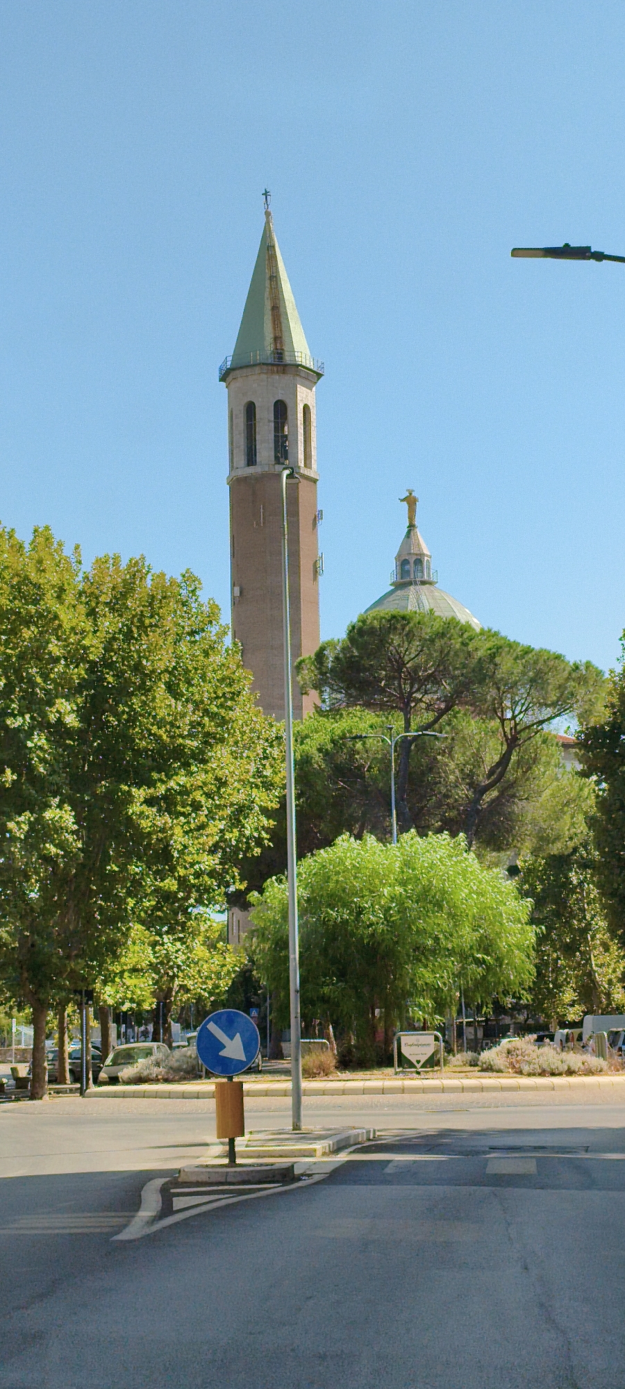 The image captures a street view in Grosseto, Italy, featuring a tall, brick bell tower topped with a green spire and a cross, adjacent to a building with a dome crowned by a statue of Christ the Redeemer. Lush green trees flank the structures, with one large deciduous tree on the left and a cluster of evergreen trees in the center and right. A street light is visible on the right, and a blue circular traffic sign with a white arrow pointing to the right is positioned near the foreground. The road surface in the foreground shows painted lines, indicating a junction or crossing. The sky is clear and blue.