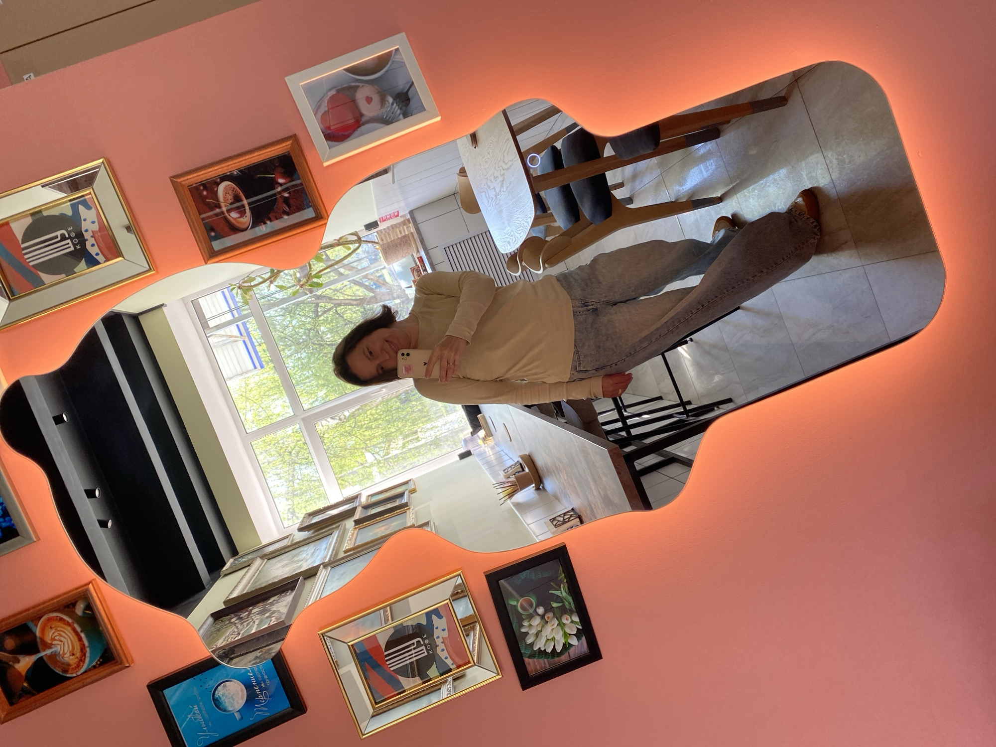 The image shows a reflection of a woman taking a selfie in a uniquely shaped mirror on a pink wall. The mirror reflects a scene of a cafe interior with tables, chairs, and a large window. The woman is wearing a long-sleeved shirt and jeans. The walls are decorated with framed pictures. The cafe appears to be in Chaikovsky, Russia, as indicated by the inscription on one of the framed pictures.