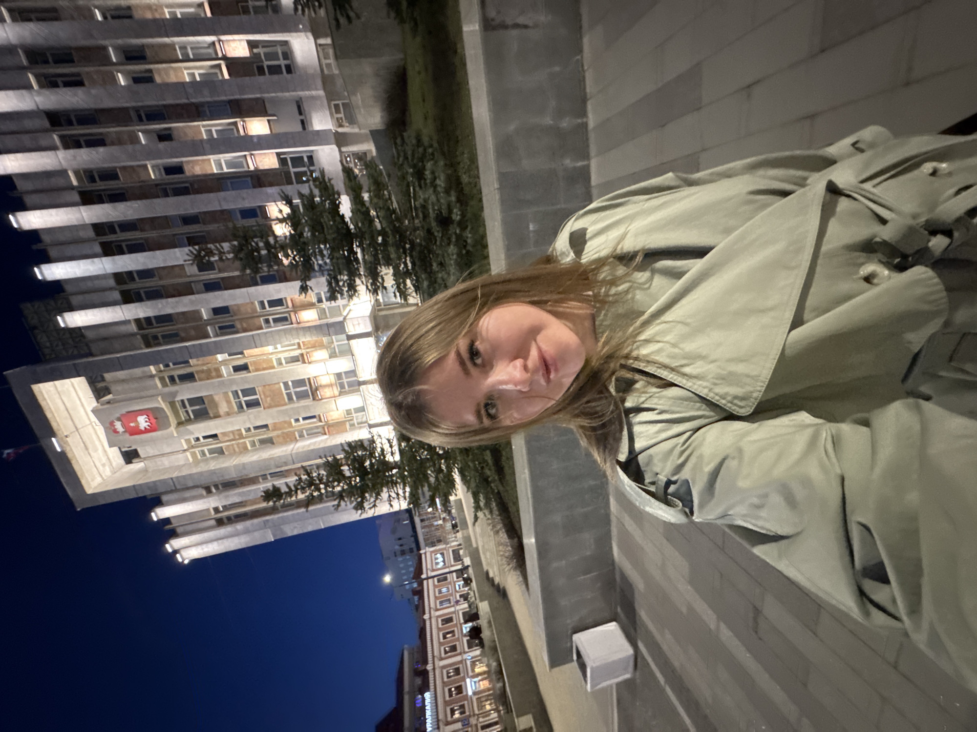 A woman wearing a green coat is visible in the foreground of the image, with the city of Perm, Russia, in the background. A tall building with many windows and a red symbol on its facade is prominent on the left side of the frame. Other buildings, with windows illuminated, are visible further in the background. The sky is dark, indicating it is nighttime. The woman is looking at the camera.