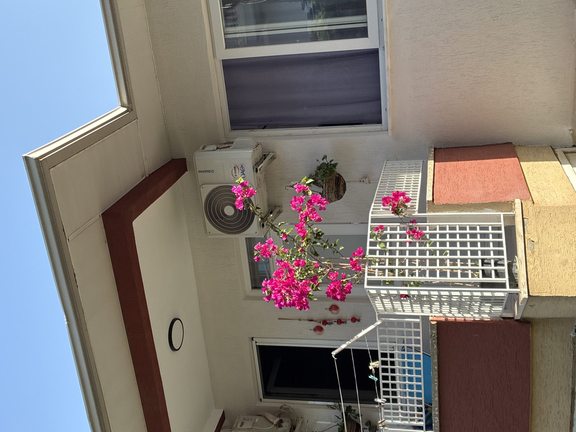 The image shows a balcony with a bougainvillea plant with bright pink flowers. The balcony has a white metal railing and a decorative white lattice screen. An air conditioning unit is mounted on the wall above the balcony. A window with a dark curtain is visible to the right. In the background, the clear blue sky is visible. This scene is likely from Alanya, Turkey, given the architectural style and the presence of bougainvillea, common in the region.