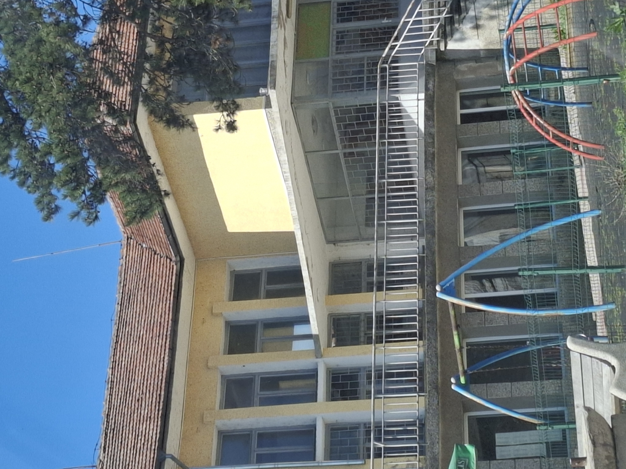 The image shows a building in Studeno Buche, Bulgaria. The building is multi-story with windows and a fire escape. There is a tiled roof on one section of the building and a yellow facade on another. A playground with colorful playground equipment is visible in front of the building. A tree is partially obscuring a portion of the building. The sky is blue.