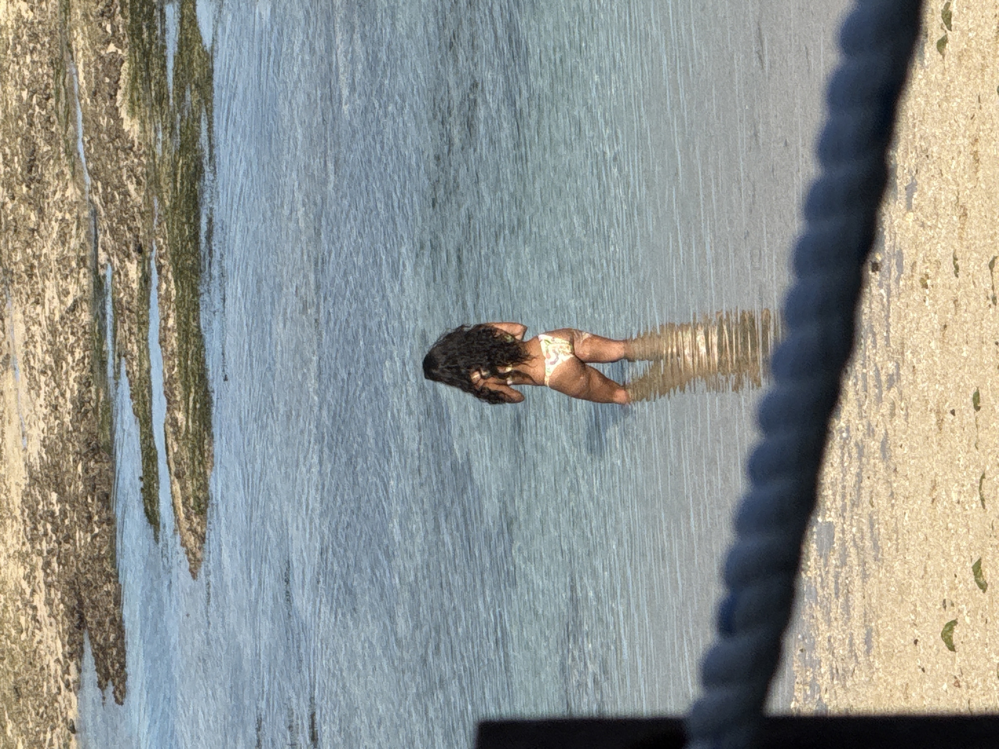 A woman with dark, curly hair is wading in the shallow water along a beach. She is wearing a light-colored bikini bottom. The water appears calm, and the beach extends on either side. Vegetation lines the left side of the image. A blurred, dark rope is visible on the right. The image was taken in Kuta Selatan, Indonesia.