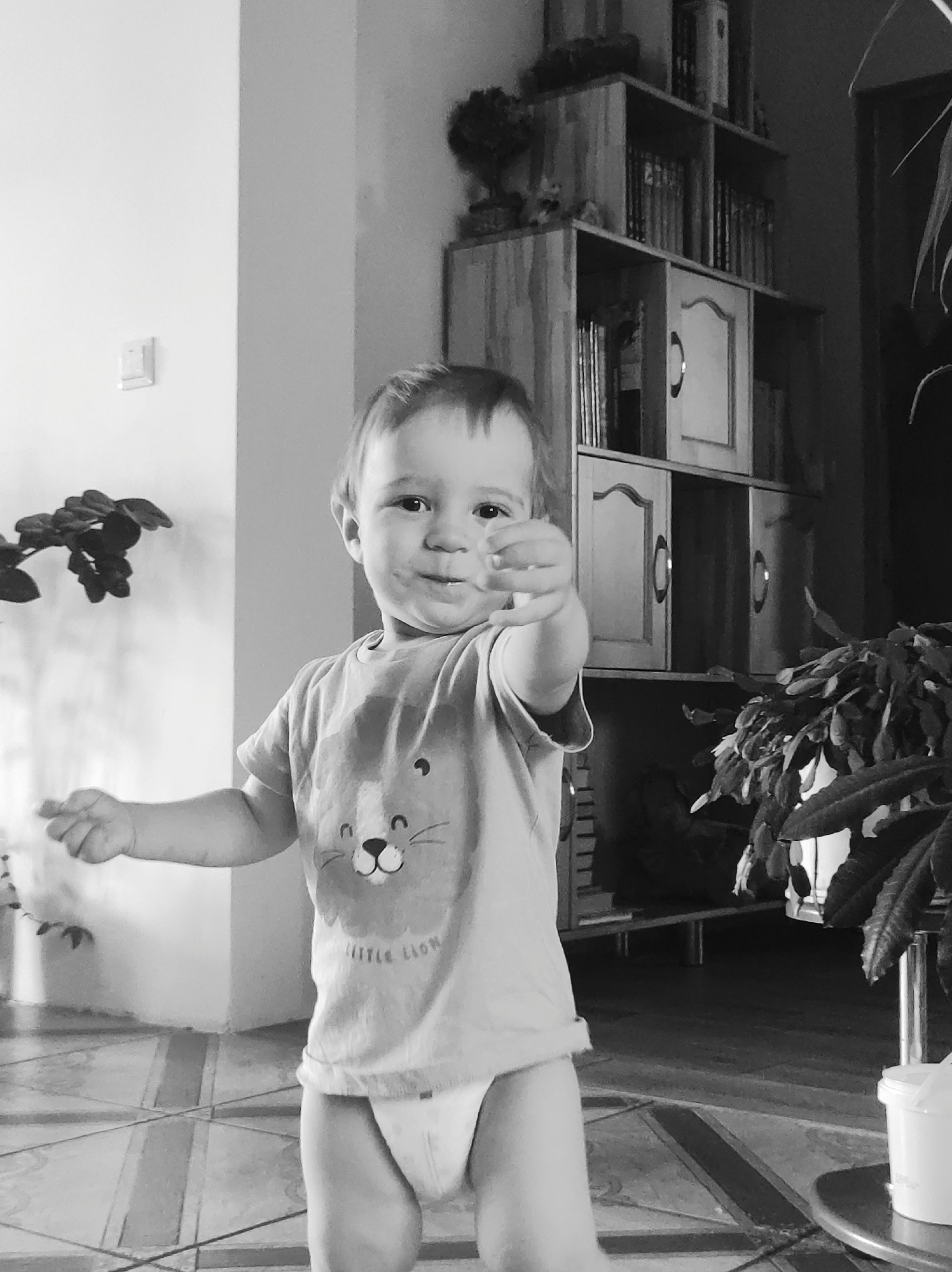 A black and white photograph shows a toddler standing indoors on a tiled floor. The child is wearing a t-shirt with a lion graphic and a diaper. The toddler's right arm is extended with the hand gesturing forward. Behind the child are a light-colored shelving unit stocked with books and decorative items, potted plants, and a wall with an electrical switch.