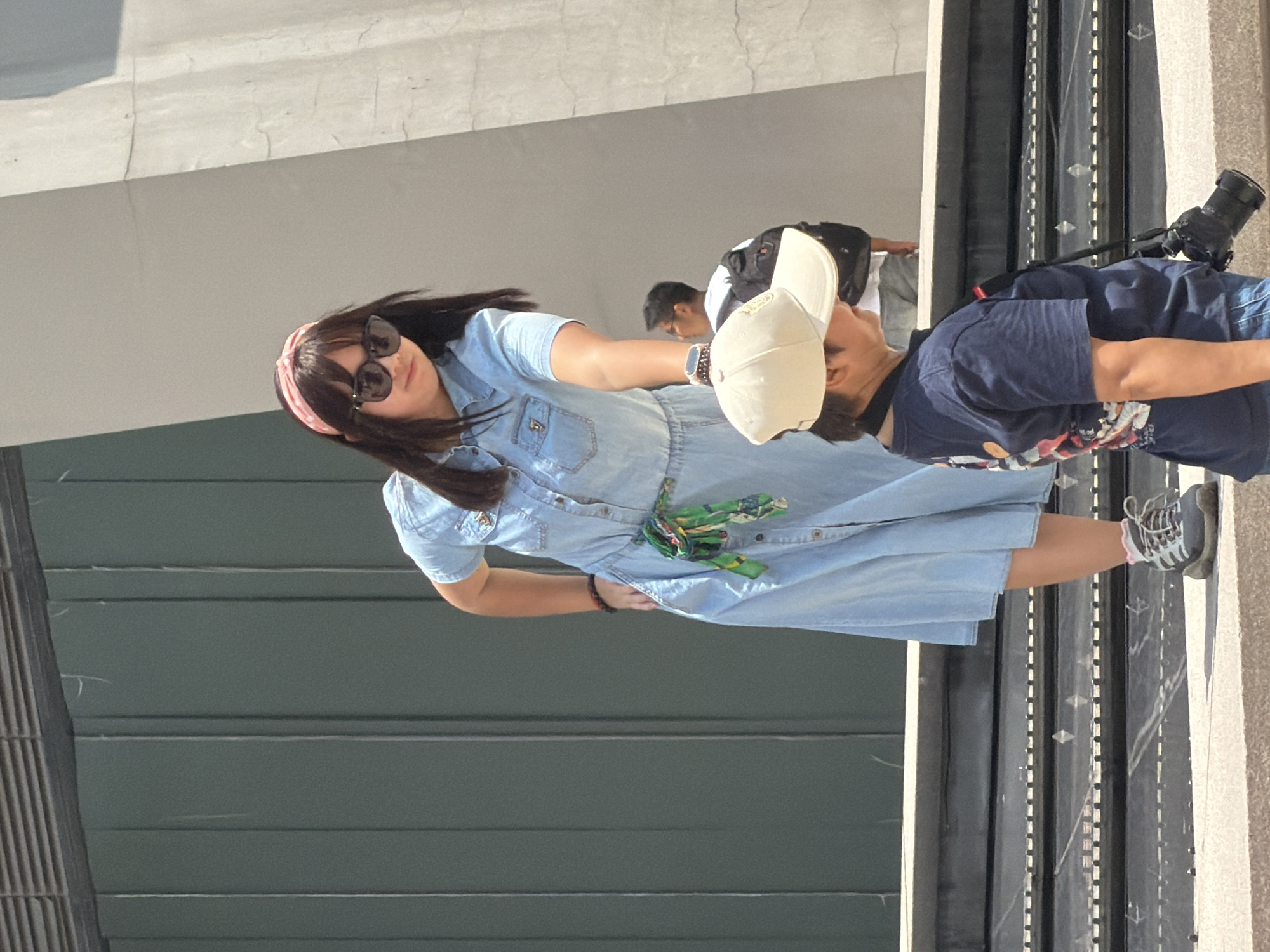 The image shows a woman in a light blue denim dress and a young person wearing a navy blue t-shirt and a light-colored baseball cap. The woman has long dark hair and is wearing sunglasses and a pink headband. She is reaching out towards the young person, who is holding a camera. The background features a large, textured, light-colored wall and a dark green wall with vertical lines. There are also steps in the foreground. The scene appears to be outdoors in a public area. The image is taken from a slightly elevated perspective.
