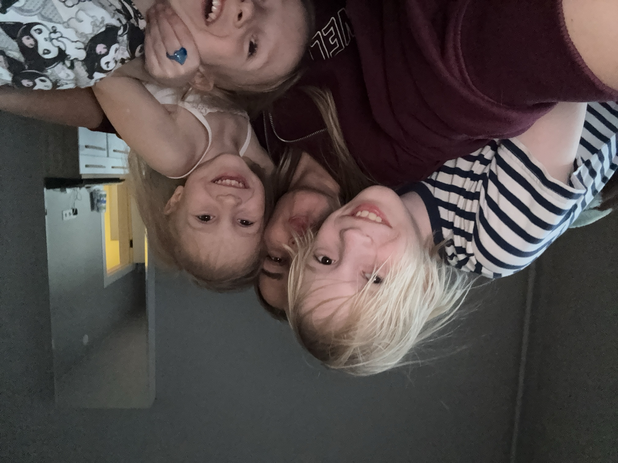 A topsy-turvy, tilted selfie captures a woman and three children, crammed together and smiling into the camera. The woman is centered, her face partially obscured by the children clustered around her. A child on the left wears a white tank top, another wears a light blue ring, and a third wears a black-and-white patterned shirt. The woman is wearing a maroon-colored sweatshirt. A child on the right wears a navy and white striped shirt. The location appears to be indoors, with a hallway visible in the background on the left side of the image. Given the instruction to locate this in Krasnoyarsk, Russia, it's impossible to pinpoint the precise address, but the interior suggests a domestic environment within the city.