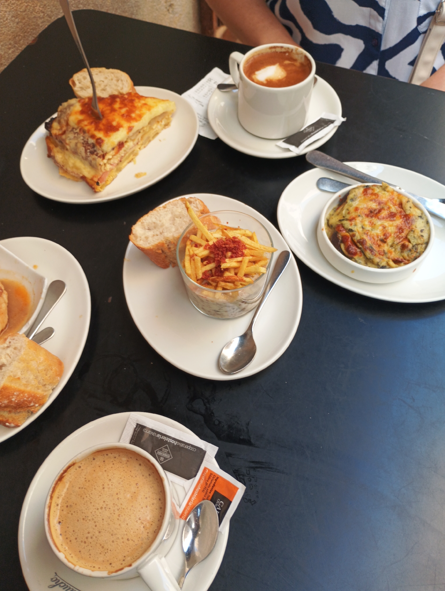 This is a top-down, medium shot photograph of a table at a cafe in Pamplona, Spain, featuring several plates of food and a cup of coffee. The table surface is dark and appears to be made of a matte material, possibly wood or laminate.
In the upper left quadrant of the image, a slice of lasagna or a similar layered baked dish is presented on a white plate. A fork is embedded in the dish, indicating it is being eaten or has just been prepared. A piece of bread is placed next to the lasagna.
Slightly to the right and below the lasagna, a white mug filled with a frothy, light brown beverage, likely coffee with milk or a latte, sits on a matching saucer. Two packets, one black and one orange, are tucked under the rim of the saucer. A silver spoon rests to the right of the saucer.
In the center of the image, a clear glass bowl contains a portion of what appears to be thinly cut fried potatoes topped with red seasoning. A piece of bread is positioned on the rim of the bowl, and a silver spoon is placed below it on a white plate.
To the right of the fried potatoes, a white ramekin holds a baked dish topped with melted cheese. This dish also appears to be a savory starter or tapa. A knife and fork are placed to the right of this ramekin on a white plate.
In the lower left quadrant, a white plate holds two pieces of bread and a small bowl containing a light-colored liquid and some bread.
The background shows a person's arm and shoulder wearing a blue and white striped shirt, suggesting they are seated at the table. The environment implies an outdoor or semi-outdoor cafe setting due to the lighting and table arrangement. The presence of specific sugar packets like "cooperativahostelerianavarra" suggests a local establishment in Navarra, the region where Pamplona is located.