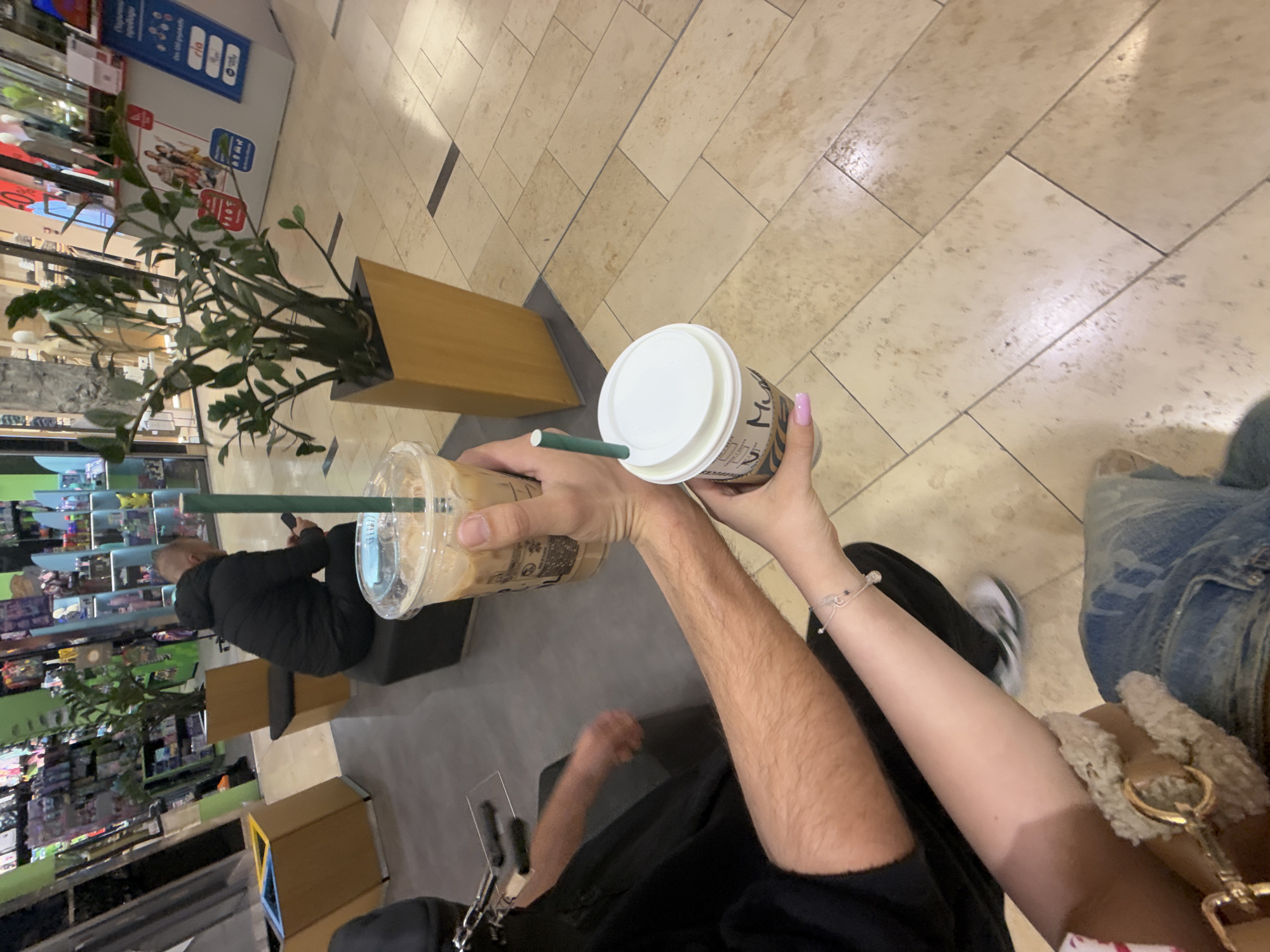 The image shows a first-person perspective of two people holding Starbucks beverages in what appears to be a shopping mall in Sofia, Bulgaria. A man is holding a Starbucks iced coffee, while a woman is holding a Starbucks hot coffee. In the background, there is a plant in a rectangular planter and a store with merchandise on display. The floor is tiled, and the setting appears to be a commercial space within a mall. The image captures the interaction between two people sharing drinks in this public place.