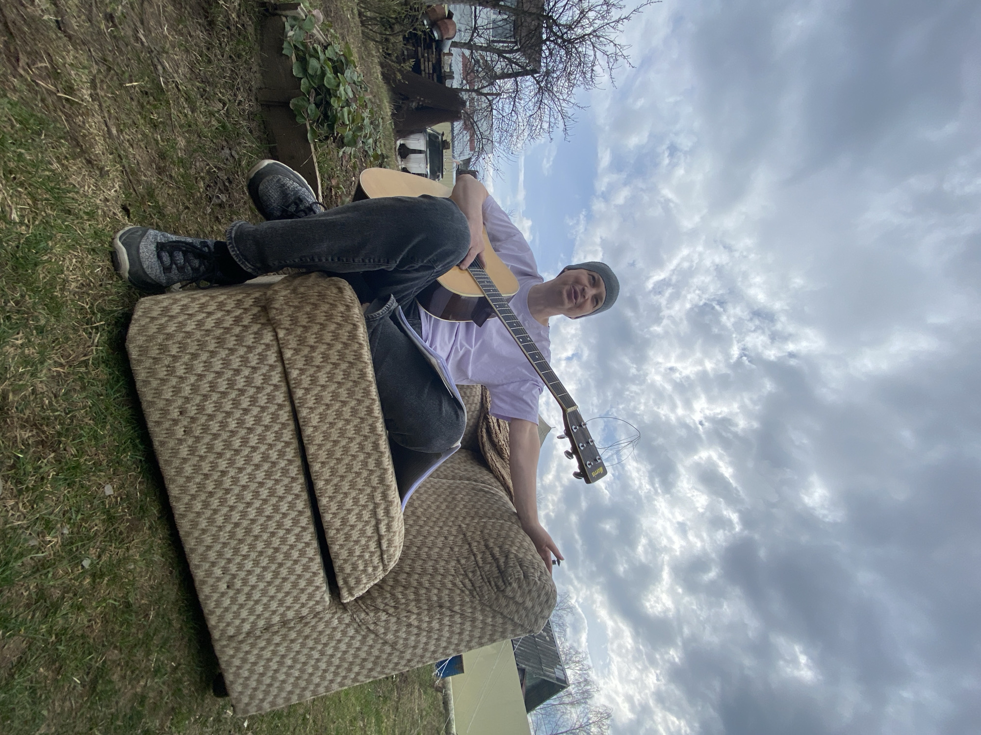 A young man sits on an armchair outside in a yard, playing an acoustic guitar. He wears a light purple t-shirt, dark gray jeans, dark sneakers, and a dark beanie. The armchair is patterned in shades of tan and brown and sits on grass. The sky is cloudy. The location is the city of Chaikovsky, Russia.
